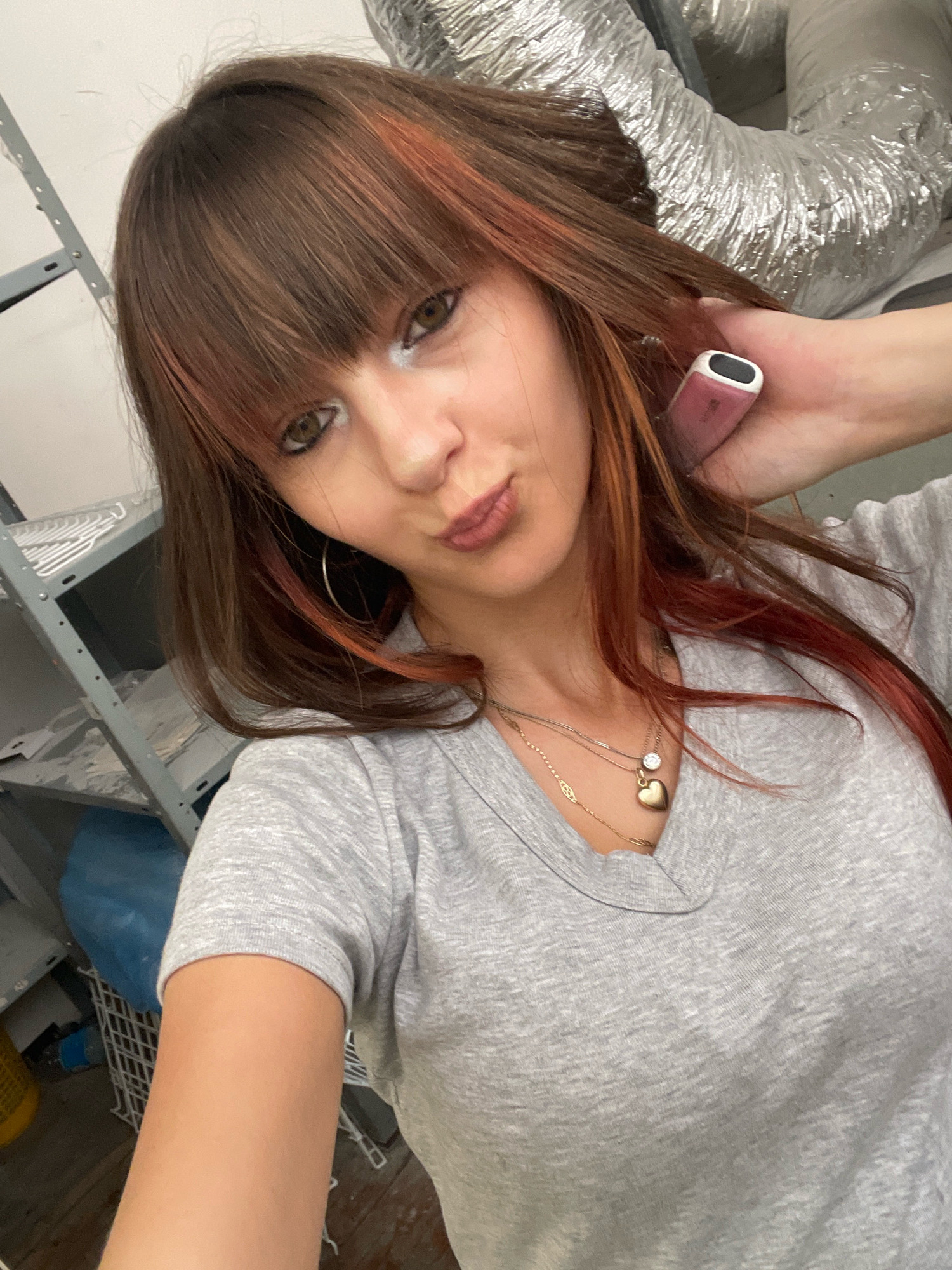 A young woman with brown hair featuring reddish-orange highlights is posing for a selfie in what appears to be a storage or workshop area. She is wearing a gray t-shirt and has on hoop earrings and a necklace with a heart-shaped pendant. Her makeup includes white eyeliner. She is holding a pink and white electronic device to her head with her right hand, and her left hand is visible as well. In the background, there are metal shelves, a blue plastic bag, and flexible metal ductwork. The scene is indoors, and based on the context provided, it is located in Raanana, Israel.