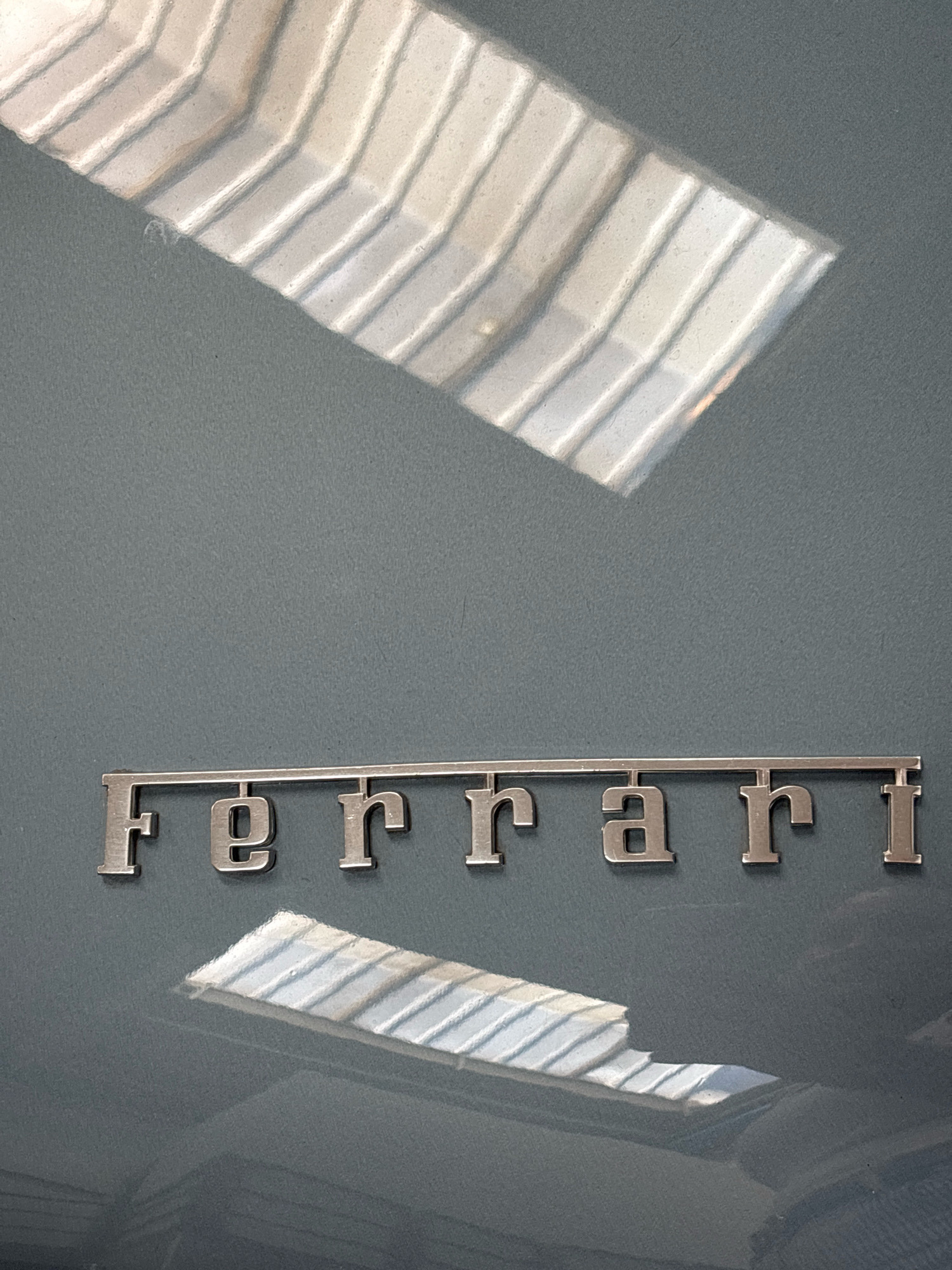 A metallic chrome emblem reading "Ferrari" is affixed to a dark grey surface. The emblem is horizontally oriented and appears to be on the exterior of a vehicle. Above and below the emblem, reflections of what appear to be a skylight or window with parallel panes are visible on the surface, creating a pattern of light and shadow. The overall scene is a close-up shot, focusing on the emblem and its surroundings. There are no people or interactions depicted. The image does not provide any information that would allow for the determination of its location in Bragni, Italy.