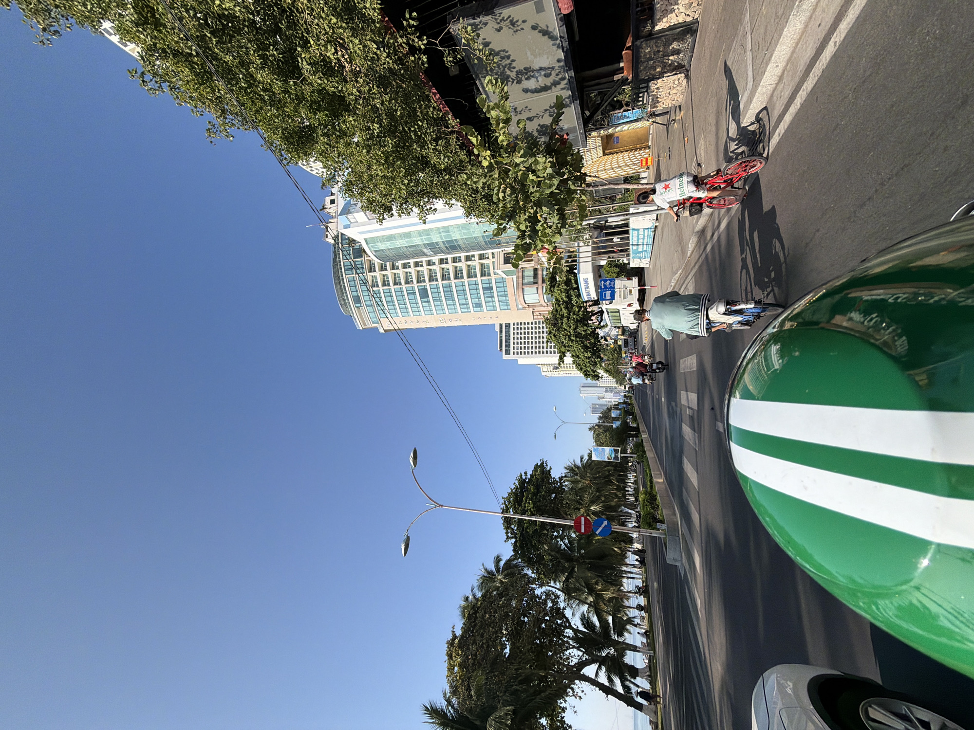 The image captures a view from a moving vehicle, likely a motorcycle, on a sunny day in Nha Trang, Vietnam. The foreground is dominated by the green and white striped helmet of the rider. In the middle ground, a paved road runs parallel to a sidewalk lined with palm trees and other vegetation. Two cyclists are visible on the road; one in a light blue shirt and the other a woman in a white t-shirt with a red bicycle. A white bus is parked on the side of the road, and several other motorcycles are also present, some carrying passengers. In the background, tall modern buildings, including what appears to be a hotel with a curved facade, line the street. Further in the distance, the ocean can be seen with a clear blue sky overhead. The scene suggests a vibrant urban coastal environment.