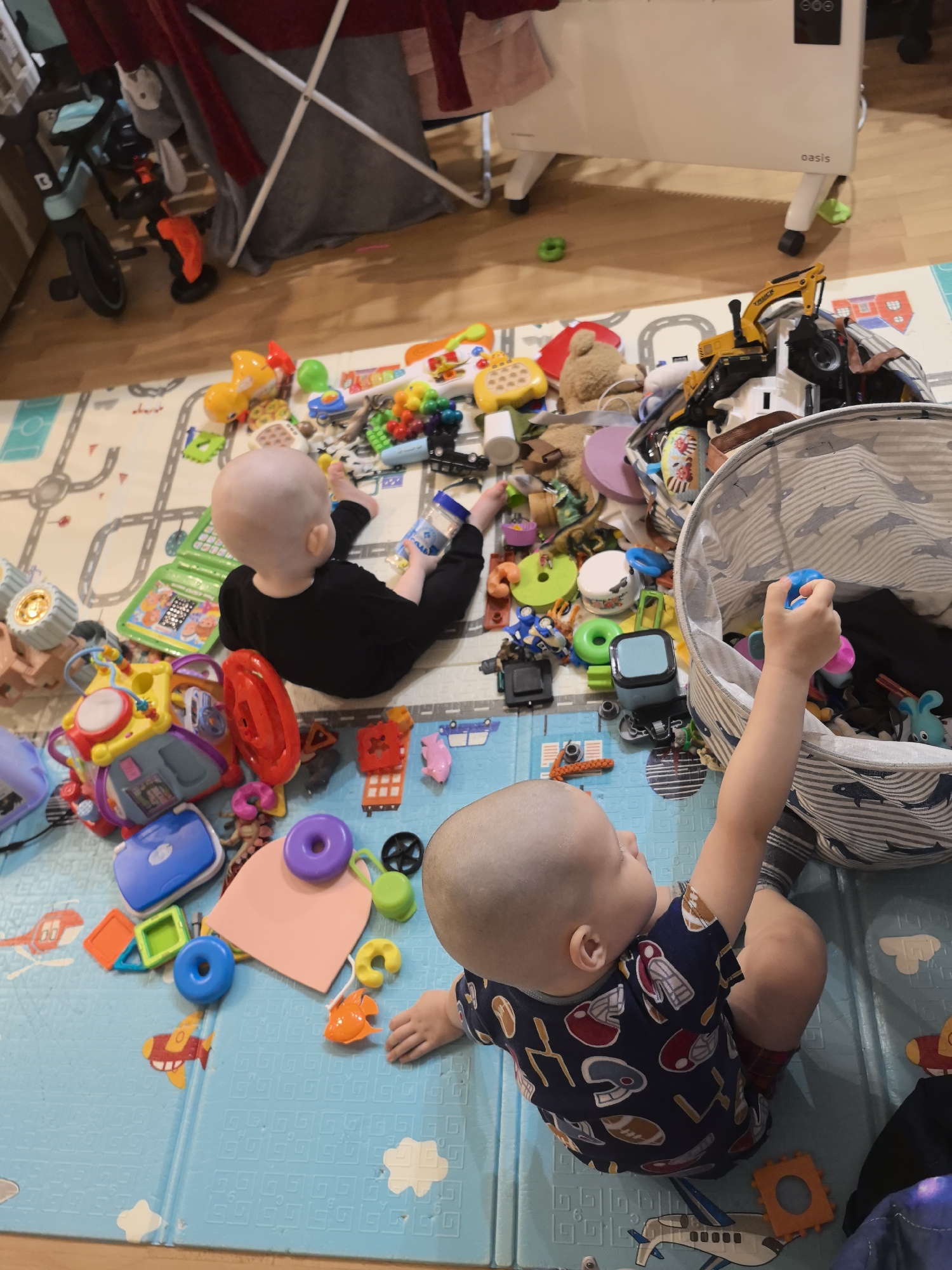 Two young children are playing on a blue and green play mat covered in toys. One child, wearing a dark long-sleeved shirt and pants, is seated with their back to the viewer amidst a pile of toys. The other child, wearing a navy blue shirt with football helmet and football motifs, is crouched near a striped toy bin and reaching inside. The play mat is patterned with roads and buildings, and the floor beneath it is light brown. In the background, there is a child's bicycle, a laundry rack, and a white electric heater. The location is an indoor play area with toys scattered on a play mat. The city of Moscow, Russia is not mentioned in the image or provided, so cannot be inferred.