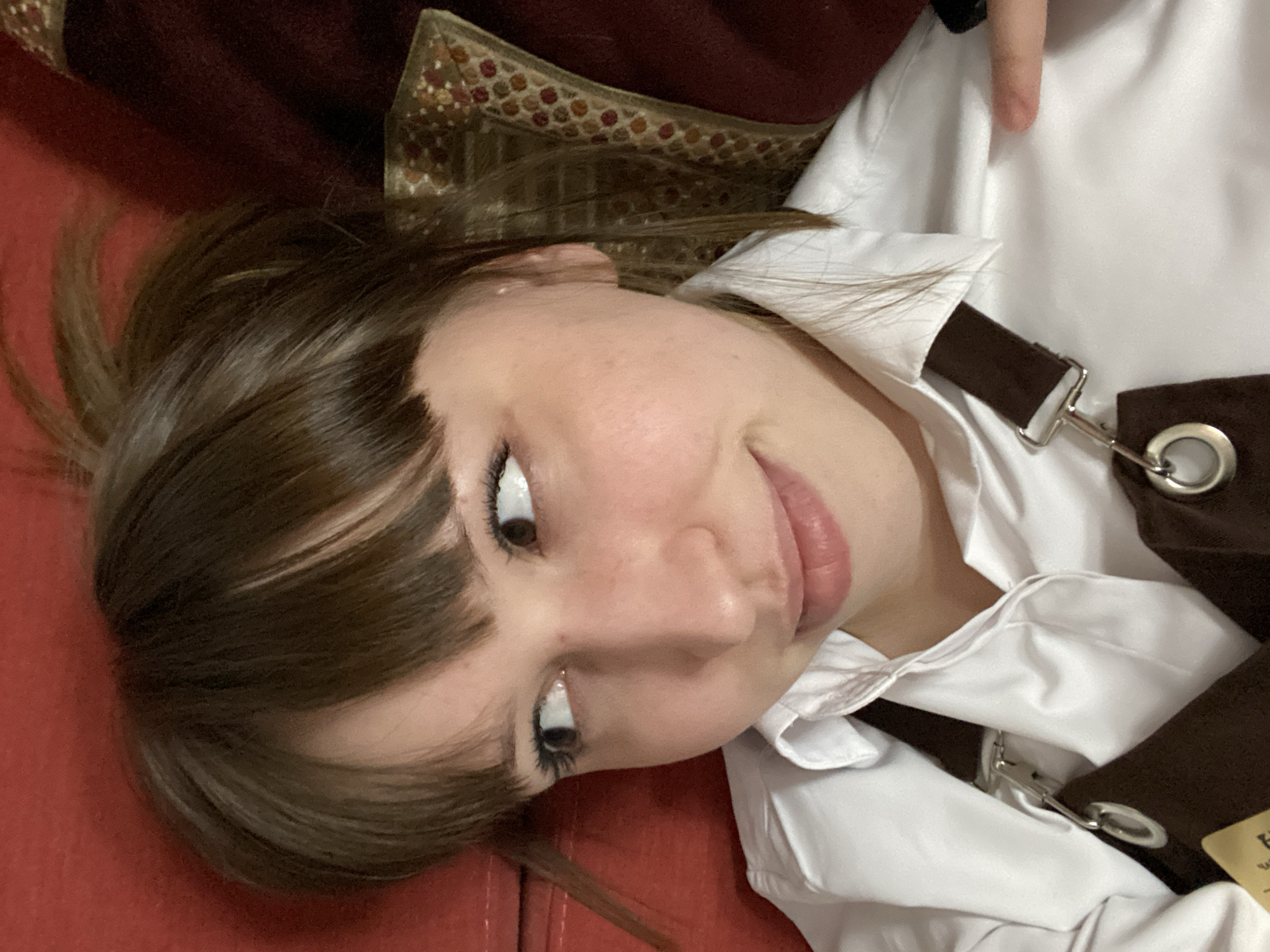 A young woman lies on a red surface, possibly a couch or bench, in a tilted close-up shot. She has fair skin, brown hair with bangs, and brown eyes looking upwards. She is wearing a white shirt under a brown garment with metal buckles and clasps. A patterned fabric is visible in the background. The precise location in Chaikovsky, Russia is not discernible from this image alone.