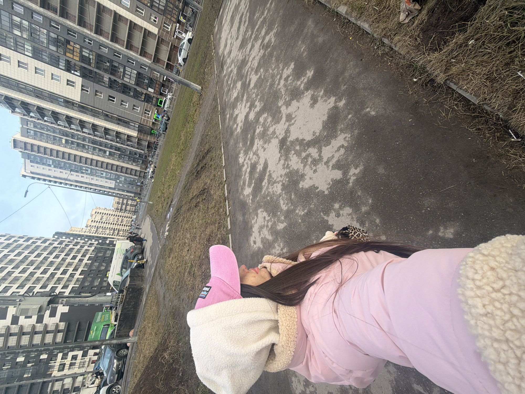 A high-angle shot depicts a person lying on a concrete walkway beside a grassy area in a city setting. The person is wearing a pink jacket with a white faux-fur lined hood and a pink hat. Long, brown hair is visible near a leopard-print hair accessory. The walkway's surface is uneven with mottled dark and light patches. Beyond the grassy area are tall, modern apartment buildings and parked vehicles. This scene is located in Saint Petersburg, Russia.
