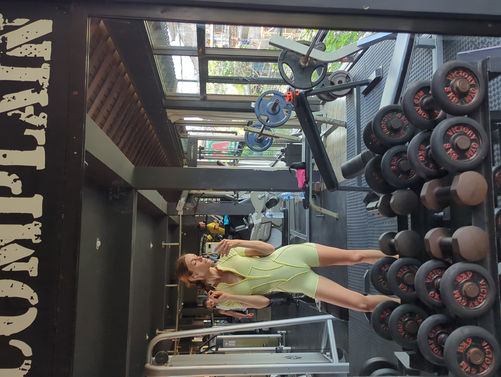 The image shows a gym interior. A woman in a light yellow romper is standing, facing to the left and holding a cell phone, possibly taking a picture or video. In the foreground, there is a rack of dumbbells. In the background, there are various pieces of gym equipment, including weight machines and bars. A window provides a view of the outside, revealing what appears to be a partially covered structure or building with some vegetation. Another person is discernible in the background. A black sign on the left with white text reading "TRAIN" can be partially viewed. The gym could possibly be located in Mengwi, Indonesia, though this detail is not directly ascertainable from the image itself.