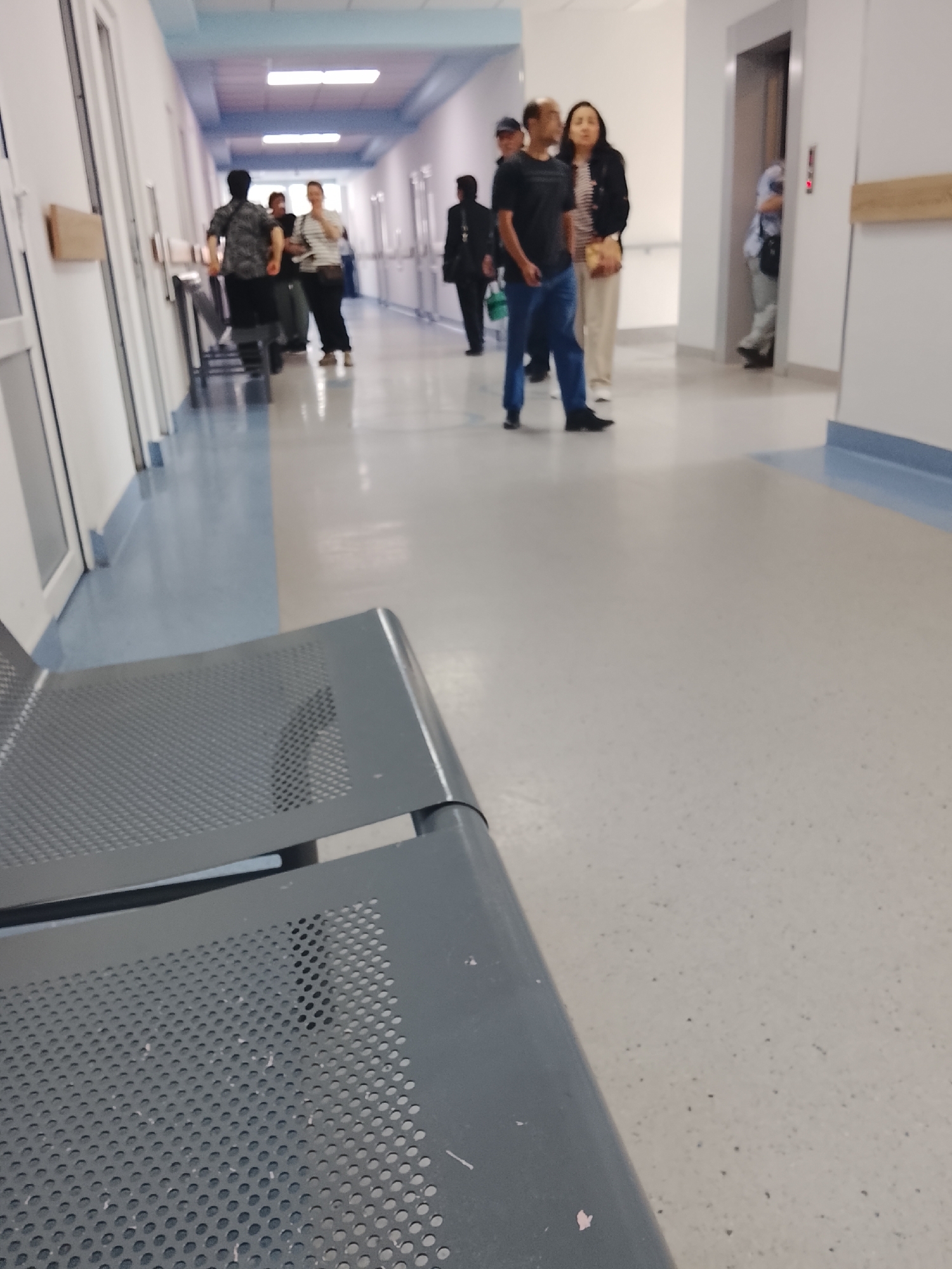 In an indoor, brightly lit corridor with a light-colored floor and white walls, a group of people are walking and standing. In the foreground, a grey metal bench with a perforated design is visible. To the right, an elevator door is open, with a person partially visible inside. Further down the corridor, several individuals are walking away from the camera. One man in the center is wearing a dark t-shirt and blue jeans. Next to him, a woman in a striped top and dark pants is holding a bag. To their left, a man in a black shirt and dark pants walks away. Another man in a black jacket is seen further down on the right. The corridor has doors on the left and a blue stripe along the bottom of the wall.