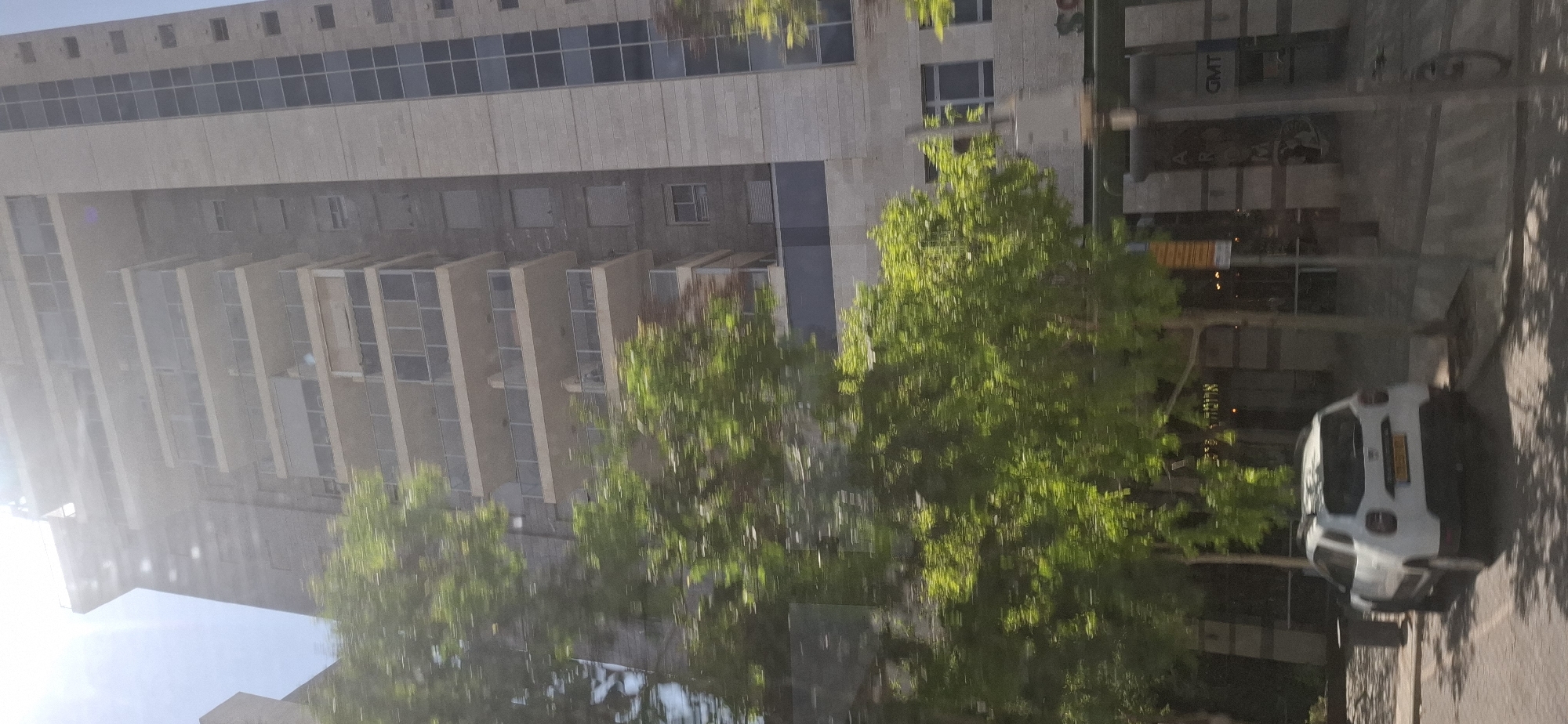 The image shows a street view in Be'er-Sheva, Israel. A white Dacia Duster car is parked on the left side of the road. In the background, there is a tall, modern building with multiple balconies and a facade of light-colored stone and dark glass. Green trees with blurred foliage line the street, indicating motion blur. The lower part of the building features storefronts with signage that includes the letters "GMT" and other illegible text. The sky is bright and appears to be daytime.