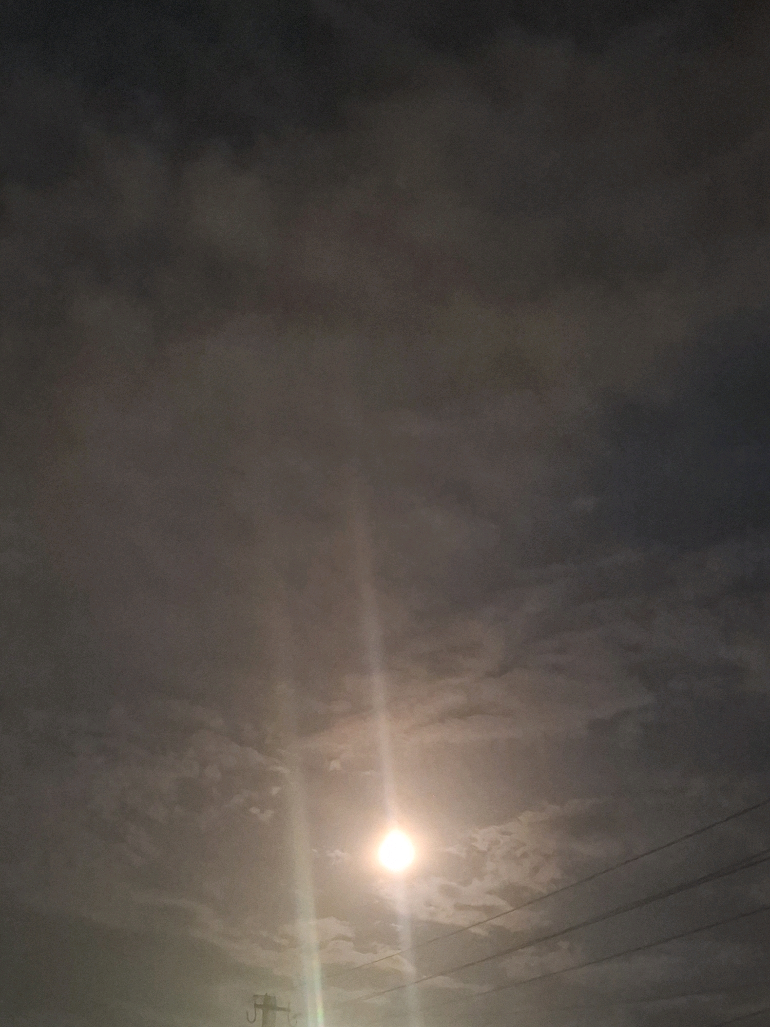 The image depicts a cloudy sky at dusk or night, with a bright light source visible towards the bottom center. This light source appears to be the moon partially obscured by clouds, or possibly a strong artificial light. There are power lines stretching across the lower right portion of the frame, and a silhouette of a utility pole is visible on the far left. The clouds are dark and heavy, suggesting overcast conditions. The scene is rendered in muted tones of gray and pale yellow from the light. No people or specific landmarks of Almaty, Kazakhstan are identifiable in this image. The environment appears to be an outdoor urban or semi-urban setting.