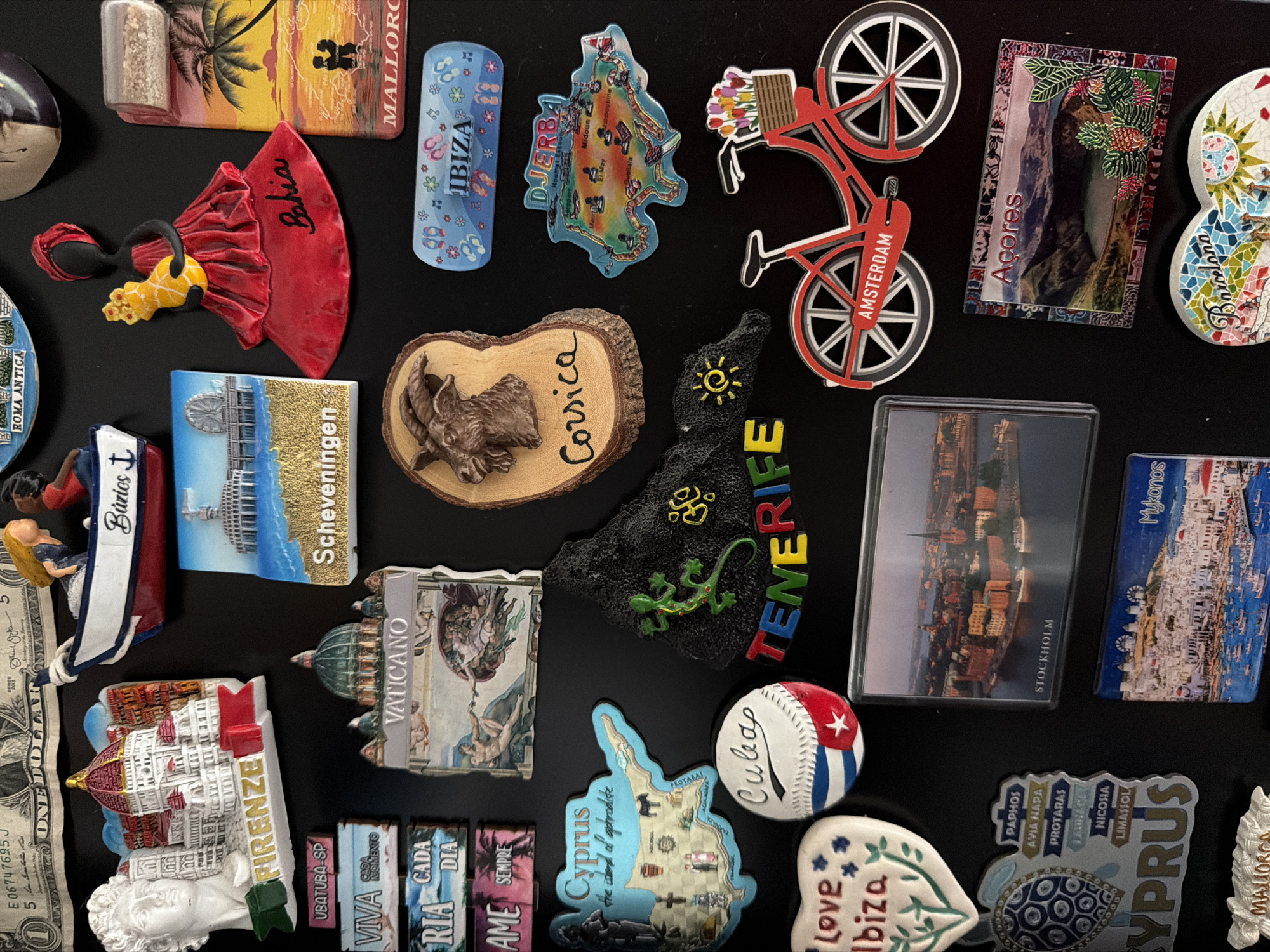 The image displays a collection of magnets on a black surface, likely a refrigerator. The magnets represent various tourist destinations and landmarks.
From the top, a portion of a US dollar bill is visible. Below that, a magnet depicts a boat with two figures. To the right, a magnet shows a woman in a red dress with "Bahia" written on it. Further right, a magnet illustrates a beach scene with palm trees and the text "Mallorca".
Moving downwards, a magnet showcases the Florence cathedral and a statue with "Firenze" written on it. Next to it is a magnet with a domed building and "Vaticano" written above an image resembling Michelangelo's "The Creation of Adam". To the right of that is a magnet with a goat's head on a wooden slice, labeled "Corsica". Below the "Vaticano" magnet is a magnet depicting a pier and a Ferris wheel, with "Scheveningen" written on it.
In the middle section, a magnet represents the island of Cyprus with landmarks and text. Below "Cyprus", a magnet shows a turtle. A baseball magnet with the Cuban flag and "Cuba" written on it is positioned to the right of the "Cyprus" magnet. A magnet with a black volcanic rock shape and a green lizard, with "Tenerife" written across it, is located below the "Vaticano" magnet. To the right of the "Tenerife" magnet is a magnet with a bicycle and the text "Amsterdam".
Towards the bottom, a heart-shaped magnet with "Love Ibiza" is visible. To its right, a rectangular magnet shows a cityscape and is labeled "Stockholm". Below "Stockholm" is a magnet depicting a coastal town, labeled "Mykonos". Another rectangular magnet to the right of "Amsterdam" displays a landscape and is labeled "Açores". At the very bottom, a mosaic heart-shaped magnet has "Barcelona" written on it.
There are no discernible elements that would place this scene specifically in Esch-sur-Alzette, Luxembourg. The magnets represent international travel destinations.