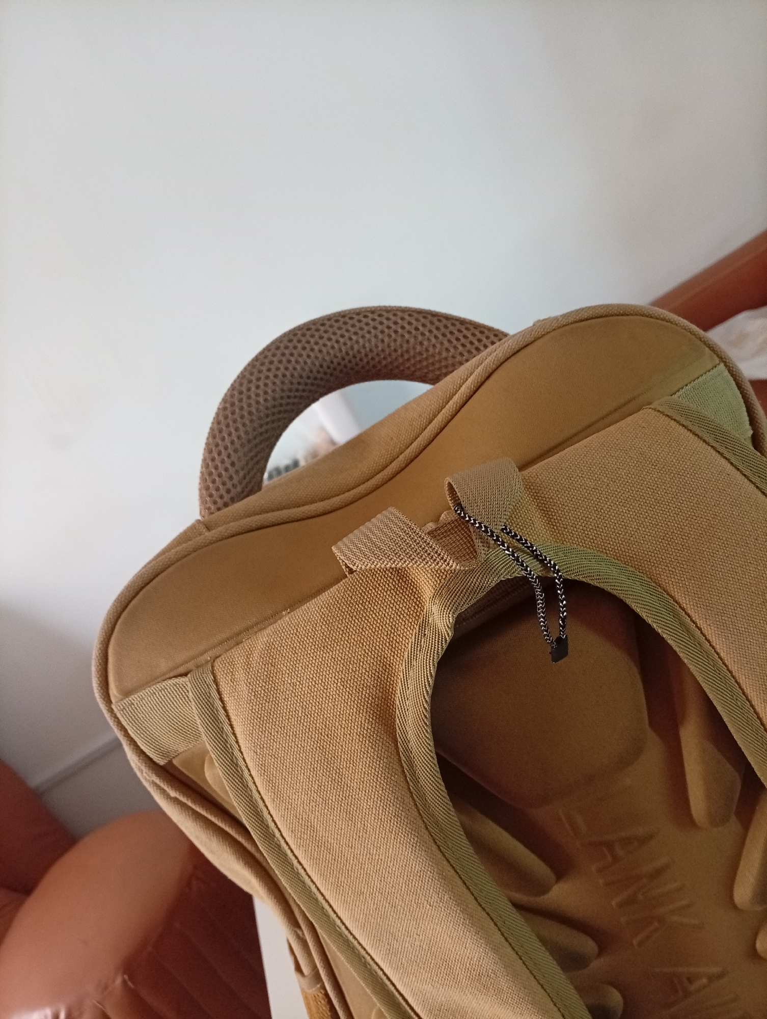 The image shows a close-up of a tan-colored backpack. It is unclear if this image was taken in Jos, Nigeria, as there is no supporting evidence of location in the image. The backpack has a padded handle and straps. A black and silver drawstring is visible on the top of the backpack.