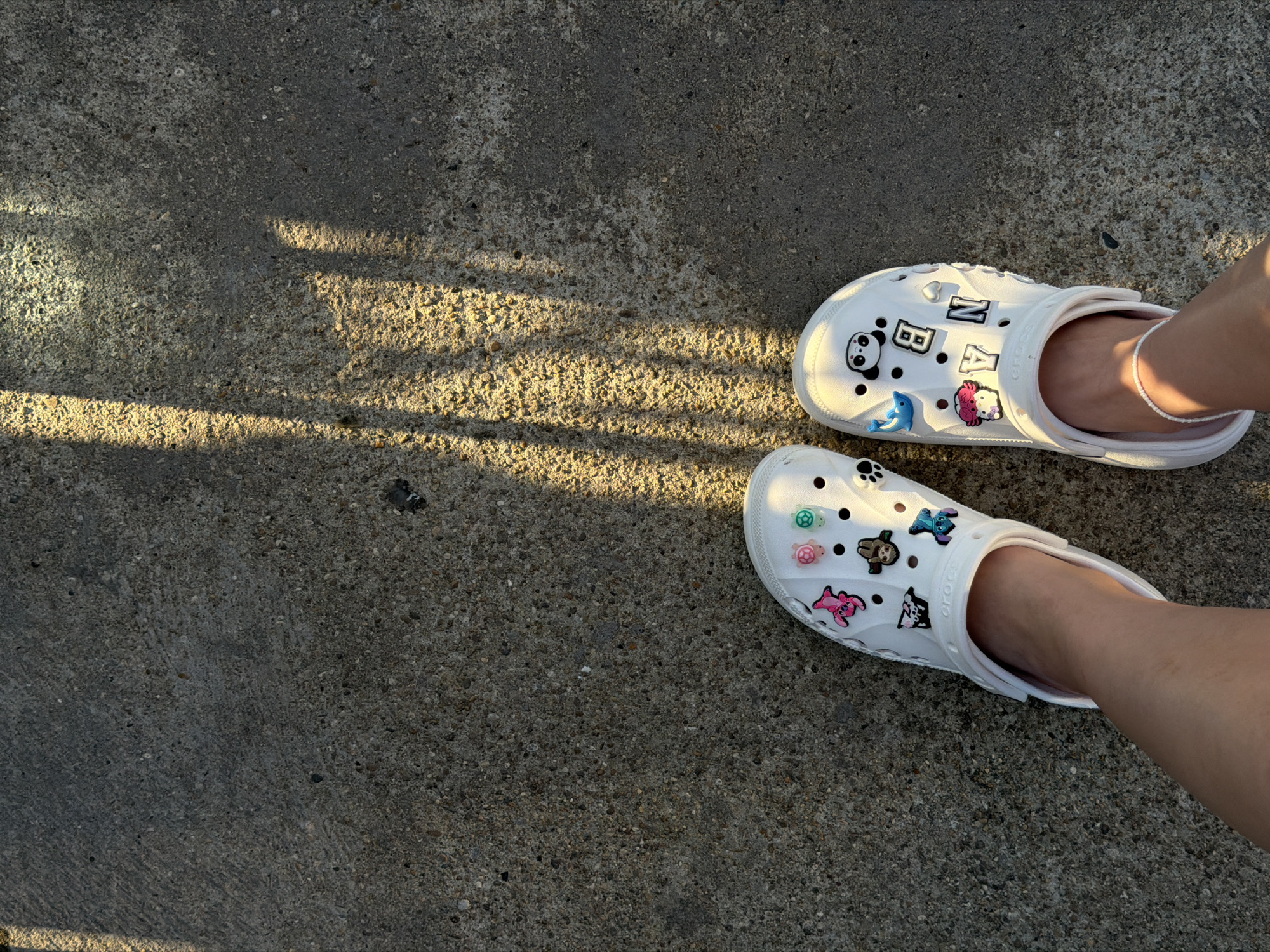 The image shows a pair of white Crocs shoes with various charms, worn by a person standing on a concrete surface. The shoes are decorated with charms including a pink pig, turtles, a panda, letters, and other cartoon figures. Sunlight casts distinct shadows across the concrete, suggesting it is outdoors. The concrete appears rough and textured, with small pebbles visible. The scene is devoid of any specific landmarks that would indicate a location in Lovech, Bulgaria, or any other city.