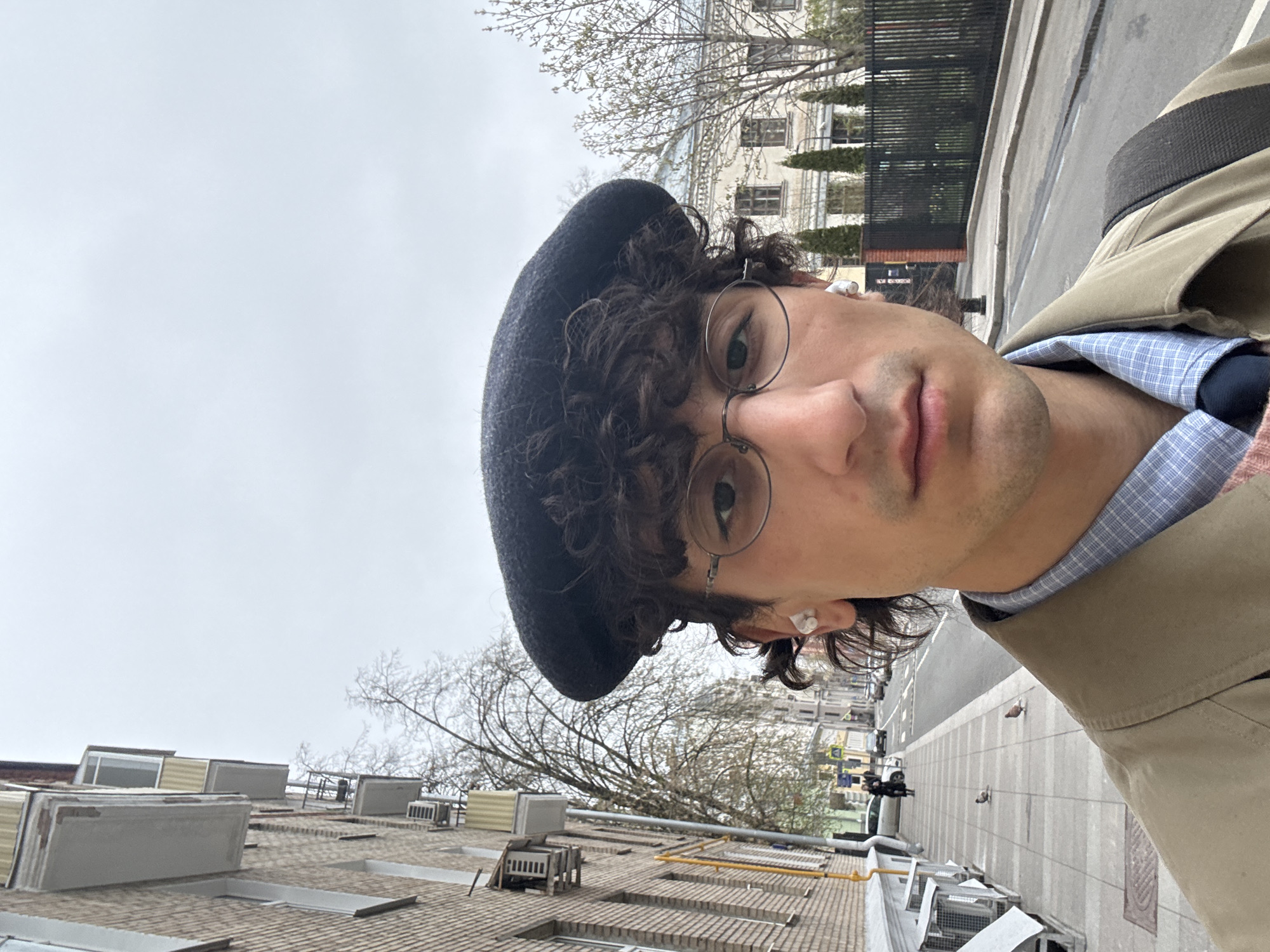 A selfie features a young man with dark, curly hair, wearing a black beret, round wire-rimmed glasses, a light blue checkered shirt, and a tan trench coat. He has white wireless earbuds in both ears and looks directly at the camera with a neutral expression.
The image is taken at a strong tilt, creating a diagonal composition. The background shows an urban landscape under an overcast, light gray sky, suggesting daytime on a cloudy day. To the man's right, parts of a light-colored building with multiple windows and bare tree branches are visible, along with a black metal fence.
Below and to the man's left, the perspective includes the rooftops of other buildings, characterized by brown shingled roofs, various skylights, and rectangular air conditioning or ventilation units. Yellow pipes are seen running along some of these rooftops. Further down, a paved street is visible with cars parked along the curb and two pigeons on the pavement. The setting depicts a city scene consistent with Moscow, Russia. No visible text is present.