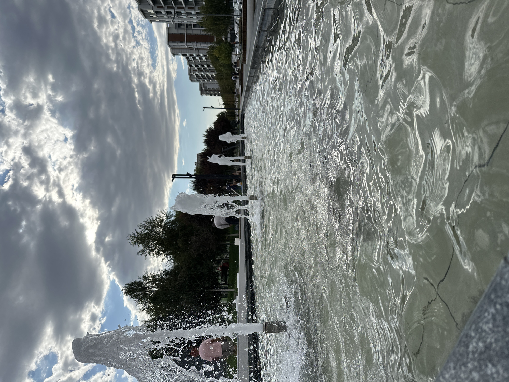 This is a medium-shot, low-angle, eye-level, horizontal exterior shot of a modern water fountain feature in the city of Tashkent, Uzbekistan. The fountain is in the foreground, with its water jets shooting upwards, creating a dynamic and slightly abstract effect against the cloudy sky. In the background, there are trees and modern apartment buildings lining a street with cars parked along the side. The sky is partly cloudy, with patches of blue sky visible between the grey clouds, suggesting it may be late afternoon or early evening. The water in the fountain is clear with ripples and reflections, and the overall impression is of a contemporary urban public space. There are a few people visible in the background, some interacting with the fountain and others walking or standing, their specific actions and interactions are not clearly discernible. The architectural style of the buildings suggests a recent development in the city.