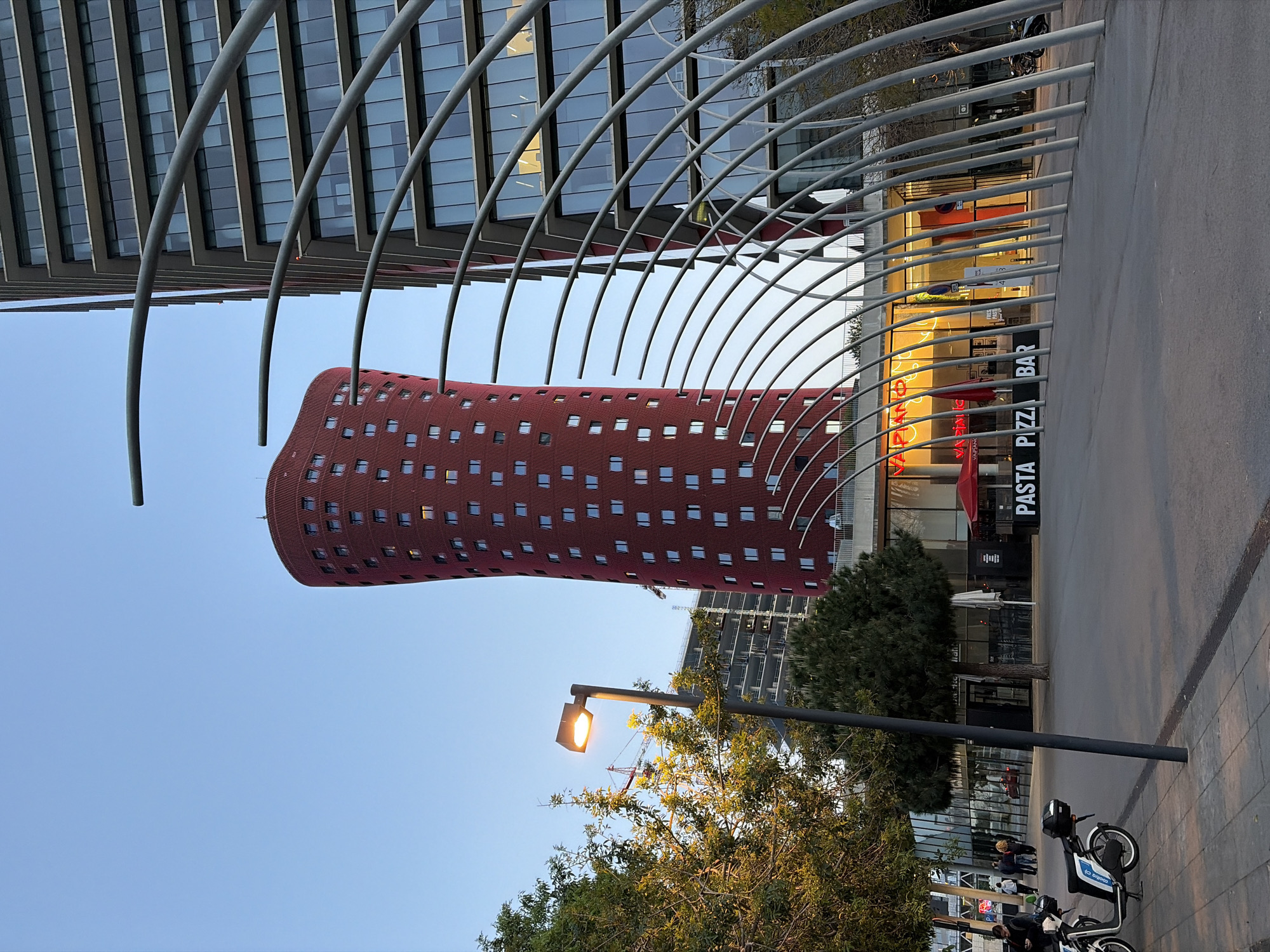 A modern urban street scene is captured at what appears to be dusk or early evening, characterized by distinctive architecture and a clear blue sky. In the foreground, a series of parallel, curving grey metal arches form a prominent, dynamic architectural canopy, framing the view.
To the left, a tall, reddish-brown skyscraper stands out with its unusual, slightly twisted or irregular cylindrical shape and numerous small, square windows. Behind the arches and to the right, another sleek, modern building with a glass facade reflects the light.
At street level, below the arches and to the right, a commercial establishment is visible. Its large windows emit a warm, golden glow. A sign clearly reads "PASTA PIZZA BAR," and the name "VAPIANO" is partially visible, indicating a restaurant. A few indistinct figures of people can be seen near the restaurant entrance.
On the sidewalk in the lower right corner, two motor scooters are parked. One has a blue container on the back with the text "O BIBOOX." A street lamp, centrally located in the mid-ground, casts a bright yellow light, further suggesting the transition from day to night. Green trees with dense foliage surround the lamp and extend into the lower left.
The overall atmosphere is peaceful and modern, showcasing a vibrant urban environment, likely in l'Hospitalet de Llobregat, Spain.