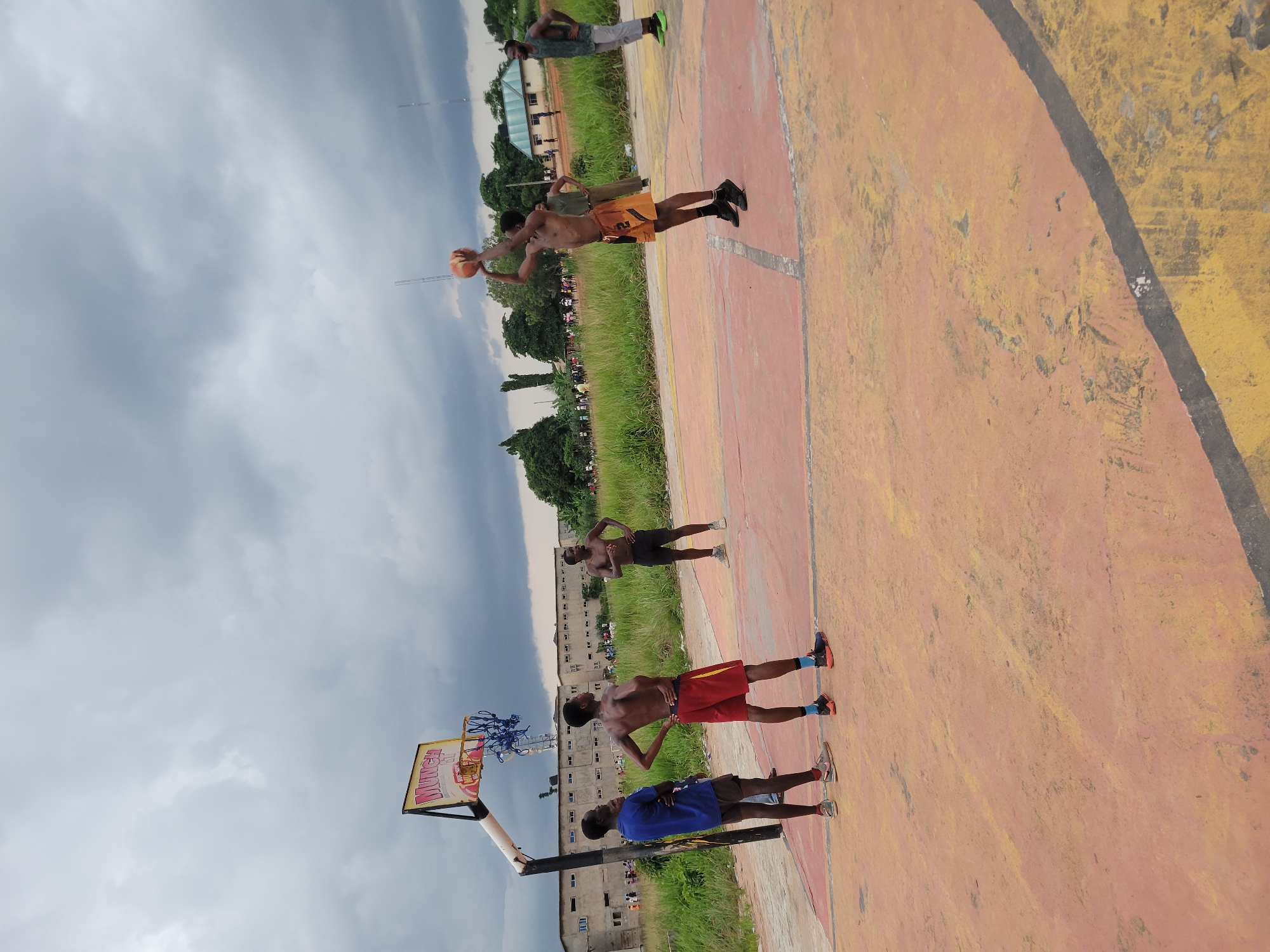In Egbagu, Nigeria, a group of young men is playing basketball on an outdoor court under a cloudy sky. The court surface is a mix of reddish-orange and yellow paint, with some wear and tear visible. A standard basketball hoop with a net is positioned on the left side of the frame.
Four individuals are clearly visible on the court. One man in the foreground, wearing a blue t-shirt and shorts, stands facing away from the basket, looking towards the game. To his right, a shirtless man in red shorts and athletic socks is also looking towards the basket with his hands on his hips. Further to the right, another shirtless man in black shorts stands with his hands on his hips. The fourth man, actively engaged in the game, is in the process of shooting a basketball. He is shirtless and wearing orange shorts with a black number "2" on them, and black athletic shoes. He is mid-jump, with the ball held high above his head.
In the background, a long, multi-story building with numerous windows is visible, suggesting an urban or semi-urban setting. Tall grass and trees line the perimeter of the court, with more distant trees and structures suggesting a developed area. Several other people are scattered in the background, some appearing to be spectators or involved in other activities. The overall atmosphere suggests a casual, community-oriented gathering centered around the basketball game.