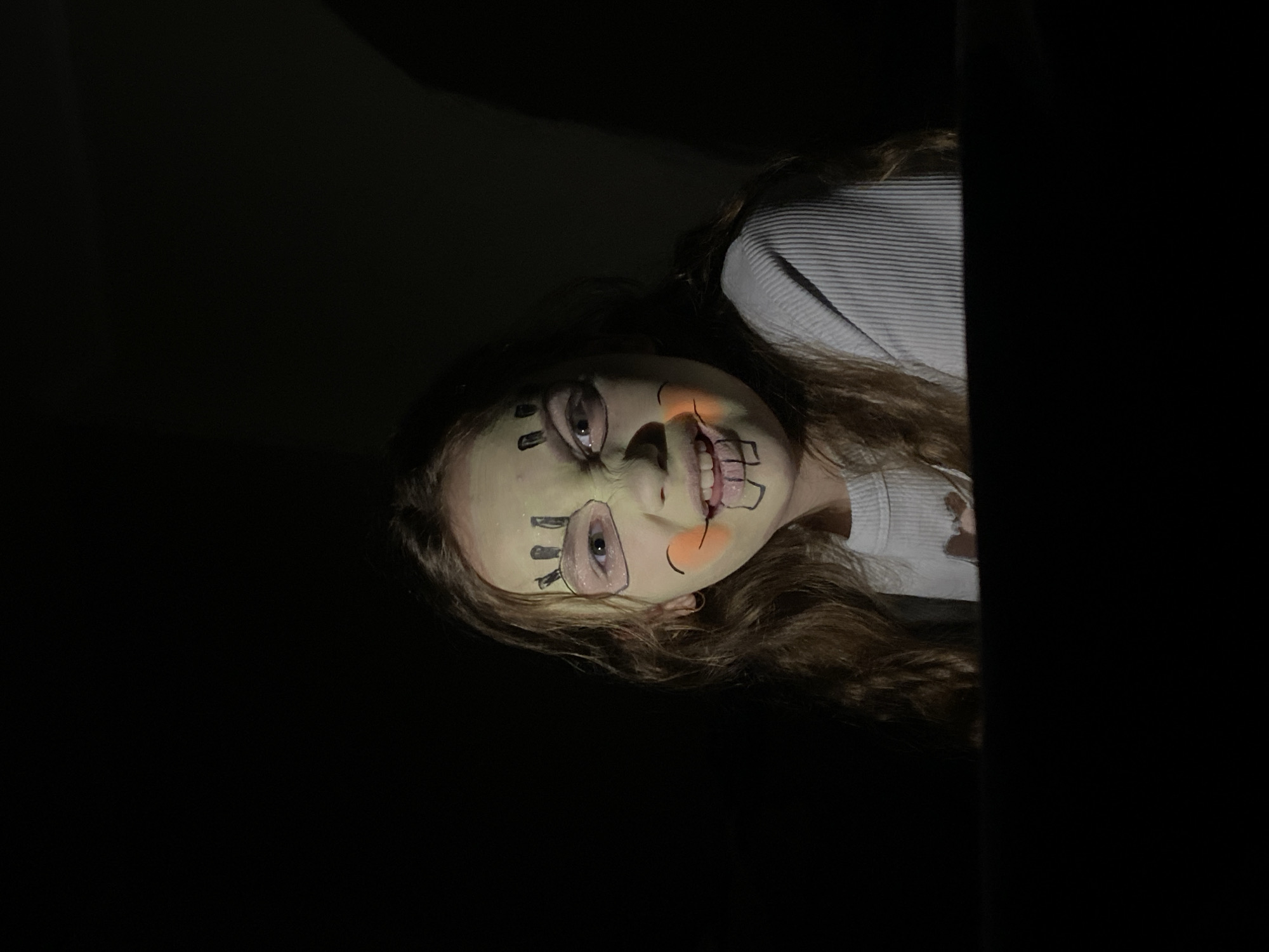 A person with pale face makeup and dark marker-like drawings on their face peeks from behind a dark, vertical edge. The person is smiling, and their hair is light brown and wavy. They wear a light-colored, ribbed top. The background is dark, suggesting a dimly lit environment. The location is Unknown, Unknown.