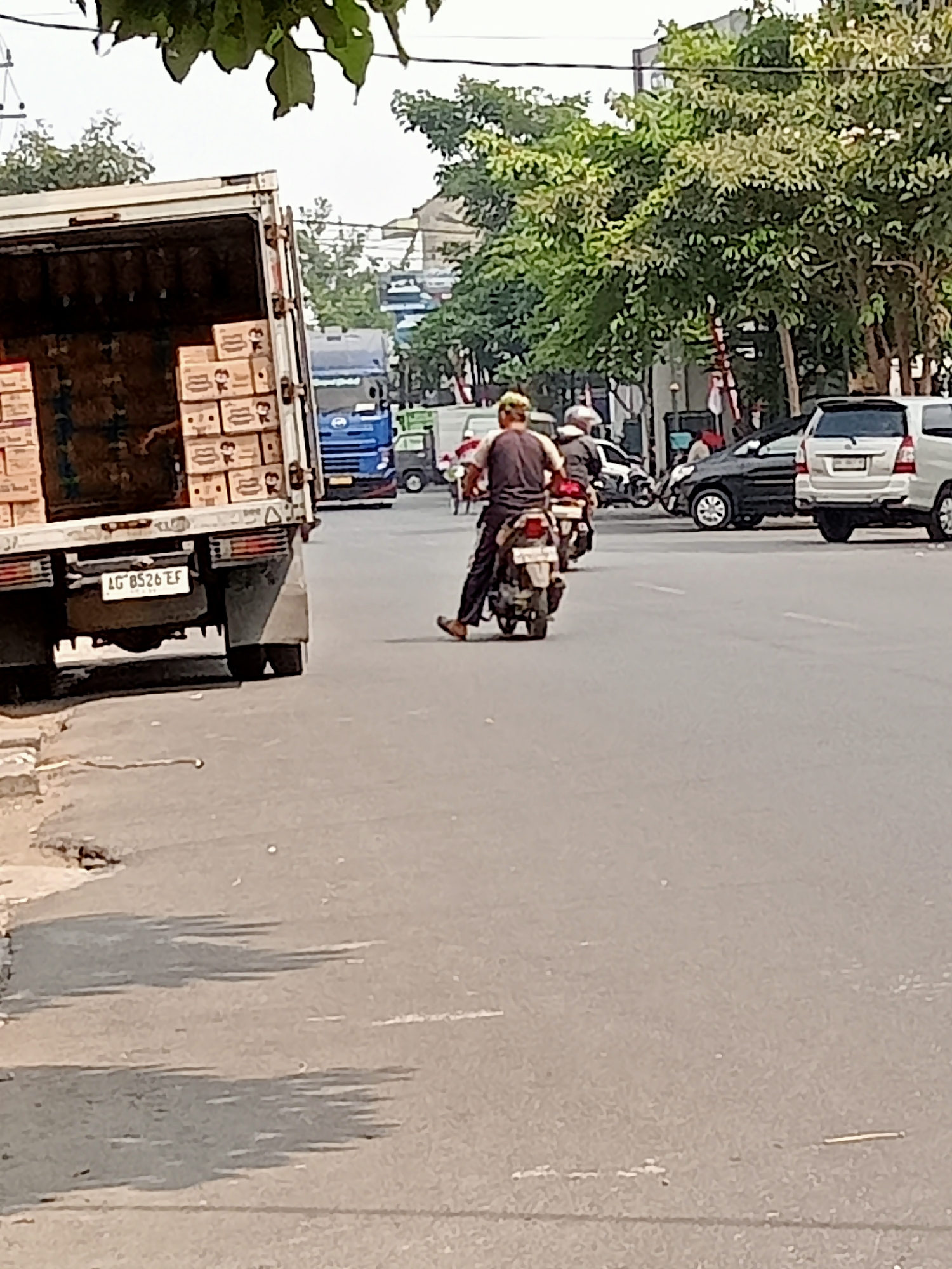In Mangunsari, Indonesia, a street scene unfolds with a large truck carrying boxes, a blue truck, and several cars. A person on a motorcycle rides away from the camera, wearing a dark shirt and pants. The environment appears to be an urban setting with trees lining the road. The truck's license plate reads "AG 8526 EF". The road surface shows signs of wear and tear.