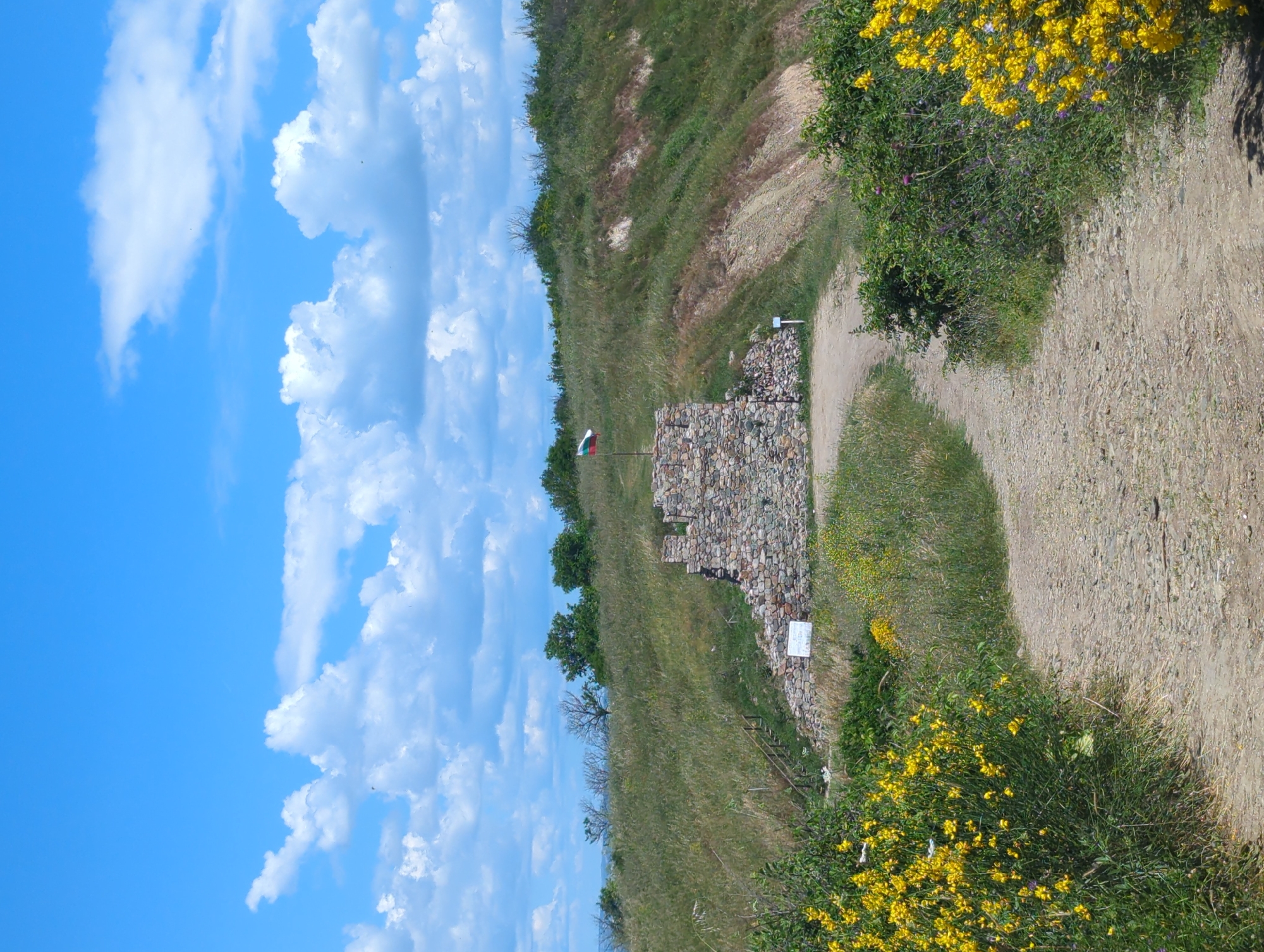 The image shows a stone ruin on a grassy hill under a blue sky with white clouds. A Bulgarian flag flies from a pole in front of the ruin. A dirt path leads up to the ruin from the foreground, which is bordered by bushes with yellow flowers. The scene appears to be outdoors in a rural or historical location.