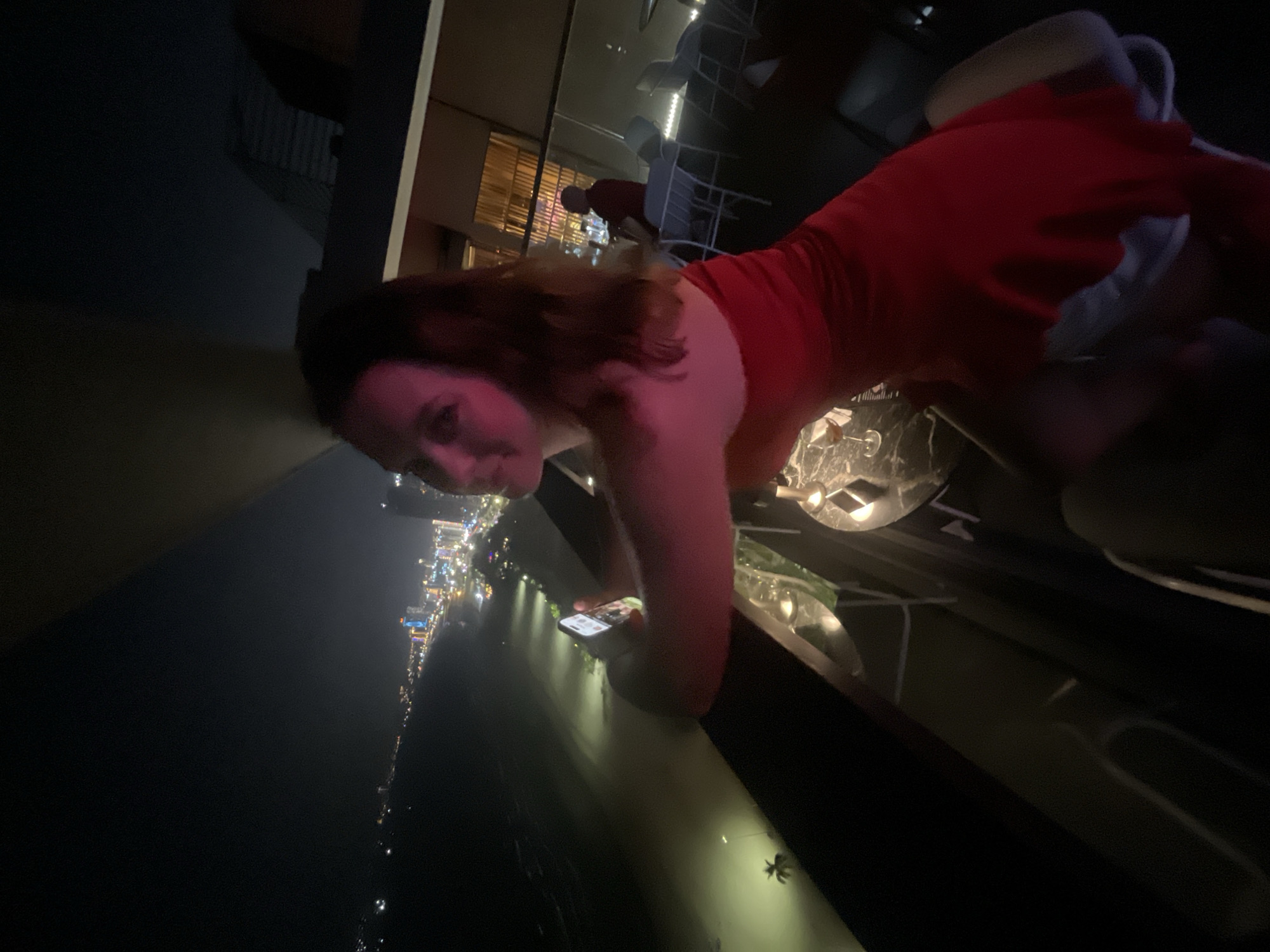 This media file captures a woman in a red dress posing for a photo from a high vantage point at night. She is leaning over a railing, with her face visible in the foreground, looking towards the camera. Her hair is reddish-brown and falls around her shoulders. In the background, there is a cityscape illuminated by numerous lights, suggesting it is nighttime. The lights stretch along what appears to be a coastline or river, with buildings and streetlights visible. There are also tables and chairs visible, indicating an outdoor seating area, possibly a restaurant or bar. The overall mood seems relaxed and perhaps a bit playful, given the pose and the context of a night out. The image is taken from a low angle, emphasizing the height and the view. The location is identified as Đà Nẵng, Vietnam.