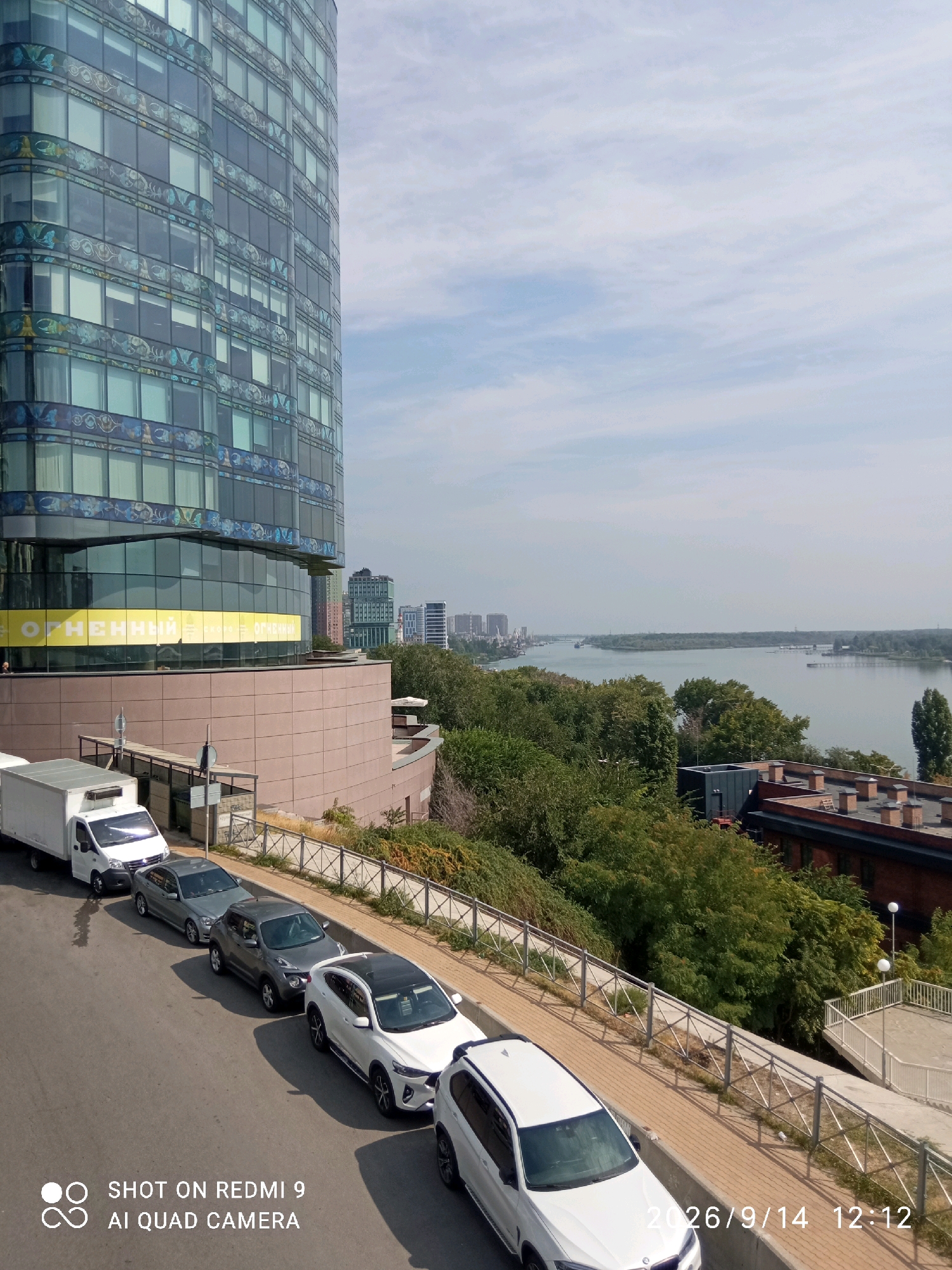 This image, taken in Rostov-on-Don, Russia, shows a cityscape with a prominent modern building on the left, featuring a glass facade with decorative elements. A wide river flows in the background, with trees and other buildings lining its banks and extending into the distance. In the foreground, a street is lined with parked cars, including a white van and several passenger vehicles. A metal fence separates the street from a slope covered with greenery and a paved walkway. The sky is partly cloudy. The timestamp "2026/9/14 12:12" and the text "SHOT ON REDMI 9 AI QUAD CAMERA" are visible in the lower right corner, indicating the image was taken with a Redmi 9 smartphone.
