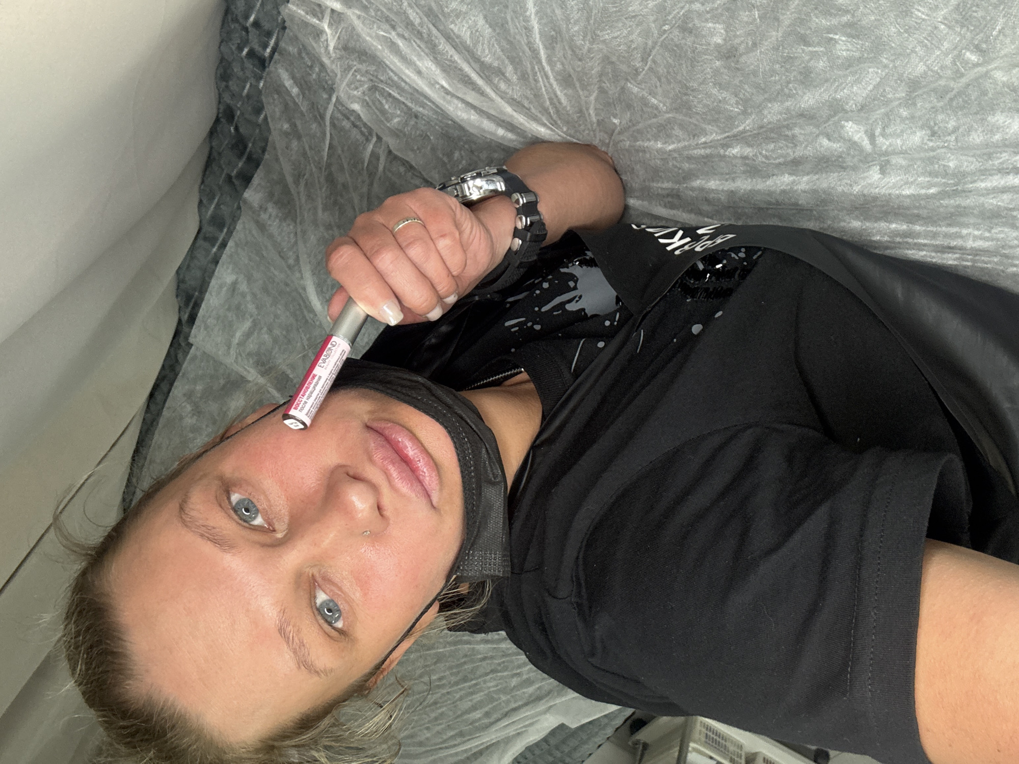 A woman with blond hair is lying on a surface covered with a wrinkled, translucent material. She is wearing a black t-shirt, a black face mask pulled down below her chin, and a watch on her left wrist. Her left hand is raised, holding a cylindrical tube with pink and red labeling. She has blue eyes and a nose piercing. The location is Perm, Russia, based on the labeling on the tube "EVABOND Восстановление после ламинирования", which uses the Cyrillic alphabet. The background includes a wall and a glimpse of what appears to be medical or salon equipment.