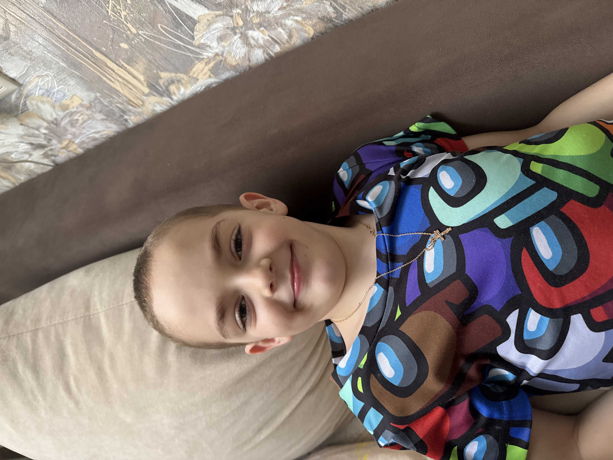 A young child, appearing to be a boy, is captured in a close-up, chest-up shot, lying on his back and looking directly at the camera. He has short, light-colored hair and is smiling gently, with a soft expression. He is wearing a colorful short-sleeved t-shirt patterned with characters resembling those from the game "Among Us," in hues of blue, red, green, purple, and brown, with white and light blue accents. A thin, gold-colored chain with a small crucifix pendant is visible around his neck.
The child is resting on a light beige or cream-colored cushion, possibly part of a sofa or bed. Behind him, to his right, is a darker brown surface, likely another part of the furniture. In the upper background, a wall or headboard is visible, decorated with a metallic-toned, abstract floral pattern in shades of gold, silver, and grey. The lighting suggests an indoor setting during the daytime. No specific activities other than resting and looking at the camera are taking place. No visible text is present in the image. The location context indicates the photo was taken in Razdolye, Russia.