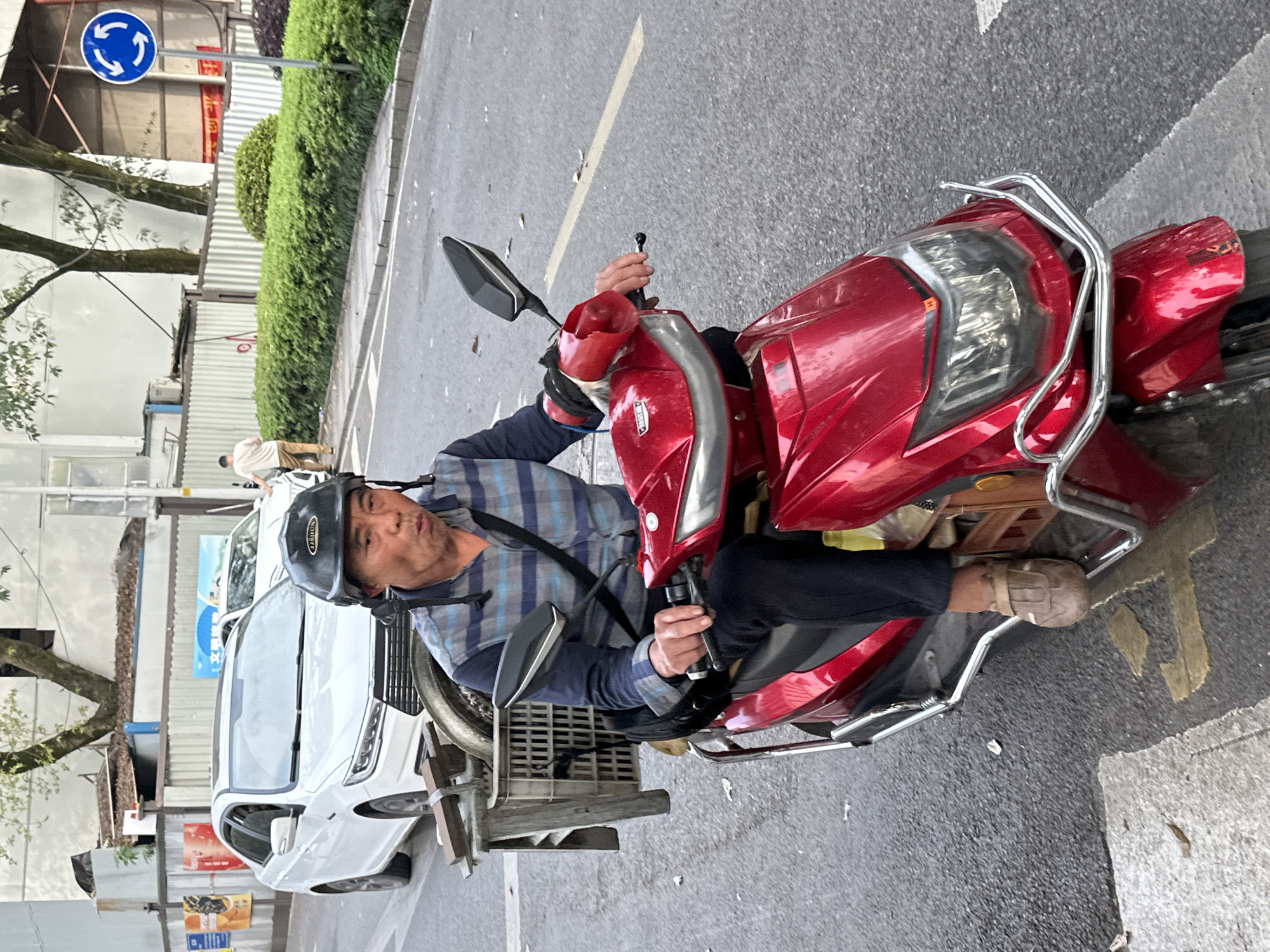 A medium shot captures a middle-aged Asian man on a red motorized scooter in a street setting, seemingly the Yuzhong District of China, evidenced by signage and architectural style in the background. He wears a black helmet, a blue plaid shirt, and dark pants, his left hand holding the handlebar with red protection. The scooter is adorned with chrome bars and a small cargo area behind the seat. To the left, a white vehicle and street signs are visible against a backdrop of trees and buildings, suggesting an urban environment. The road shows visible yellow lane markings.