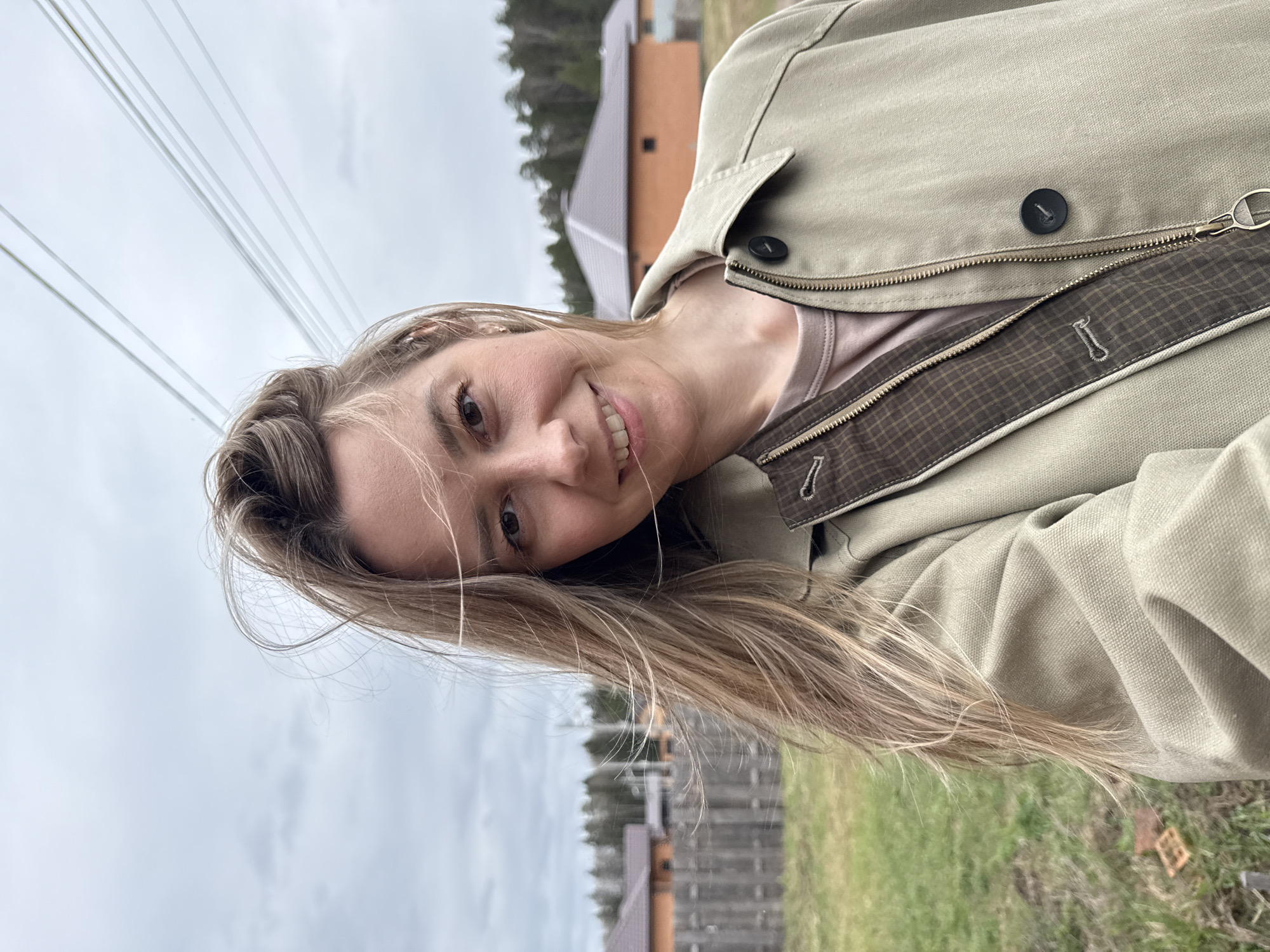 A woman smiles towards the camera; she is wearing a khaki jacket. Background shows a landscape with houses, trees, and power lines, and a cloudy sky. Location is in or near Chaikovsky, Russia.