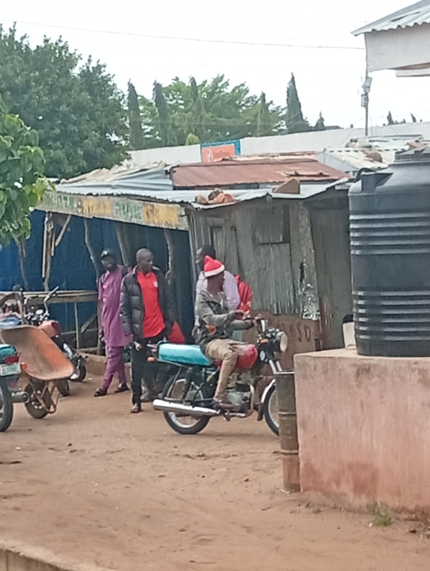 In Tumfure, Nigeria, a street scene unfolds. Several individuals are present. One man, wearing a red shirt and a black jacket, stands near a building. Another man in a purple traditional outfit is nearby, his hand on the shoulder of the man in red. Further to the right, a person wearing a red Santa hat is seated on a turquoise motorcycle, holding onto the handlebars. Another motorcycle is parked to the left, with an orange wheelbarrow partially obscuring it. Behind these individuals is a corrugated metal structure with various signs. A large black water tank is visible to the far right, next to a reddish-brown wall. The ground is dusty earth. Trees and foliage are visible in the background.
