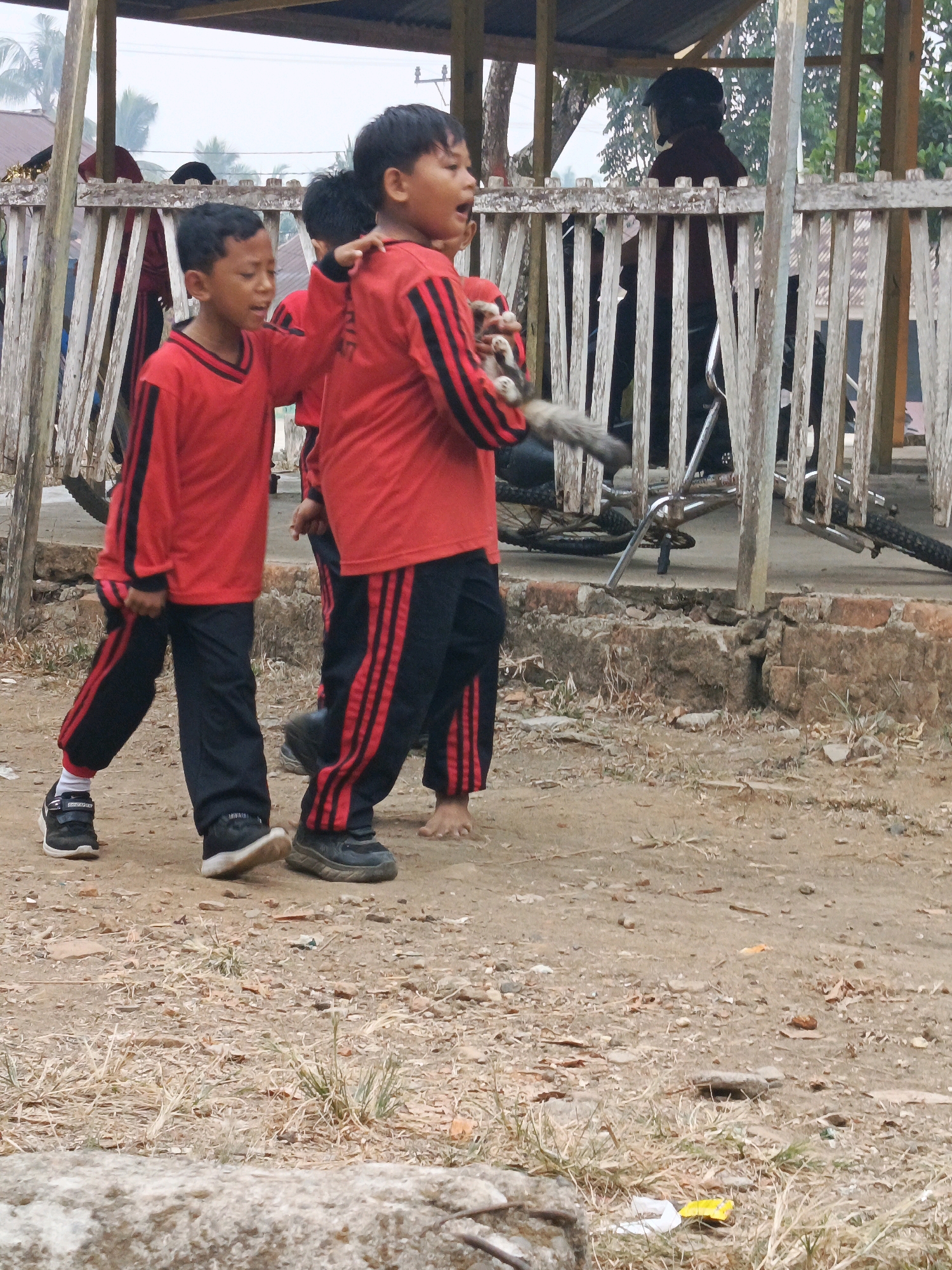 In Megang Sakti II, Indonesia, three young boys are captured in an outdoor scene. Two of the boys, wearing matching red long-sleeved shirts with black stripes on the sleeves and black pants with red stripes down the sides, are actively engaged in movement. One boy in the foreground is looking to the left, while the boy next to him is facing forward with his mouth open, seemingly speaking or yelling. A third boy, mostly obscured, is also present, wearing similar attire. Behind them, a person wearing a black helmet is seated on a motorcycle within a wooden-framed structure that appears to be an open-air shelter. The ground is covered with dirt, dry grass, and scattered debris. Two bicycles are partially visible within the shelter. The overall environment suggests a casual outdoor gathering or play area.