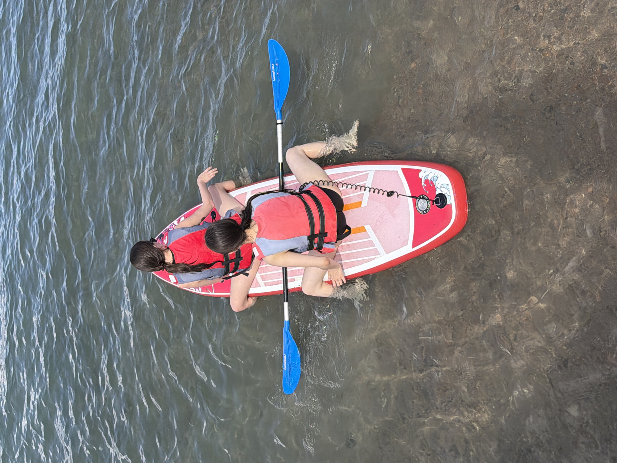 An aerial view captures two individuals on a paddleboard in shallow, clear water. Both subjects are wearing red life vests over grey shirts. They are positioned towards the rear of the paddleboard, which is primarily red with white accents and a design of waves. One person is reaching out to the side, while the other is seated. A blue paddle rests in the water next to the paddleboard, its shaft extending upwards. The water is calm and appears to be relatively shallow, with the sandy or rocky bottom visible through the water. The lighting suggests it is daytime. The overall scene conveys a sense of a recreational activity, possibly paddleboarding or kayaking, on a pleasant day.