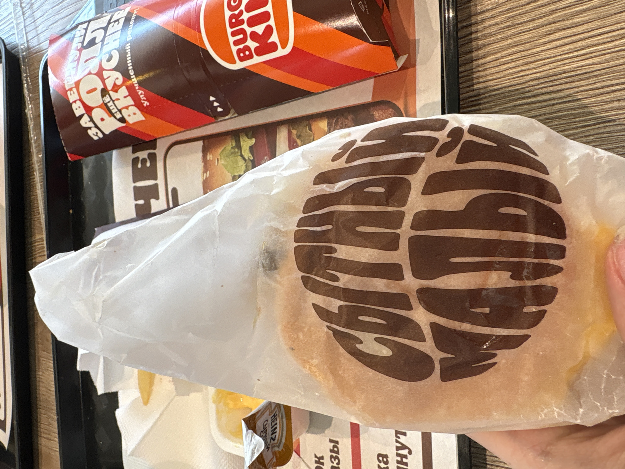 The image shows a Burger King meal, including a burger in a wrapper with the Burger King logo, a drink, and possibly fries or other sides. The location is likely a Burger King restaurant, and based on the Cyrillic text on the packaging, it is specifically in the city of Perm, Russia. A hand is visible holding the burger. The setting appears to be a typical fast-food restaurant environment. The objects include fast-food packaging and the items are all likely associated with a fast-food meal from Burger King.