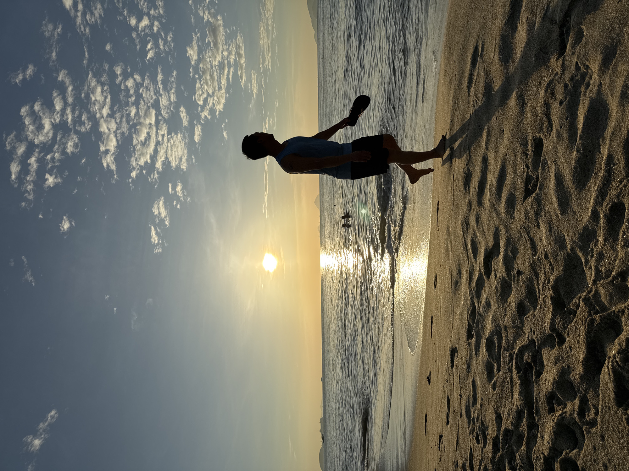 A lone man walks along the sandy shore of a beach in Nha Trang, Vietnam, during sunset. The sun, a bright orb on the horizon, casts a shimmering golden path across the gentle waves of the ocean. The sky is a soft gradient of blue and yellow, dotted with scattered white clouds. The man, silhouetted against the bright sky and sea, is wearing a sleeveless shirt and shorts, and carries a flip-flop in his right hand. His shadow stretches long across the textured sand, which is imprinted with numerous footprints. In the distance, the dark shapes of ships and islands are visible on the water. Two figures can be seen wading in the ocean further out. The scene evokes a sense of peaceful solitude and the beauty of a coastal evening.
