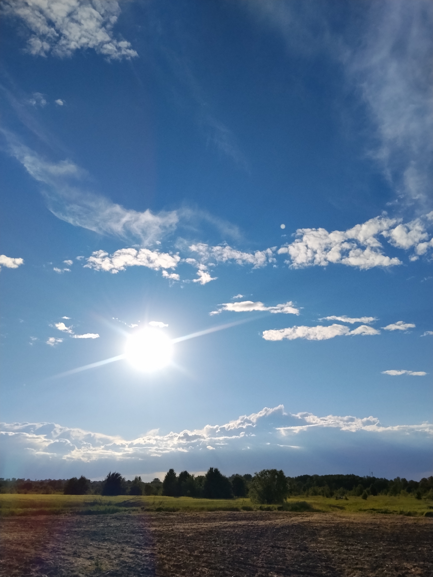 The image depicts an outdoor scene with a bright sun in a blue sky, partially obscured by wispy white clouds. A faint circular object, possibly the moon, is visible in the upper right portion of the sky. The sun casts bright rays and lens flare across the sky and towards the horizon. Below the sky, a flat landscape features a dark, plowed field in the foreground. Beyond the field, a strip of green vegetation with scattered trees and bushes forms the middle ground. The horizon is lined with a dense array of trees. The overall impression is a sunny day in a rural setting, possibly Ustyvytsya, Ukraine, characterized by open fields and woodlands under a dynamic sky. There are no people or animals visible in the image.