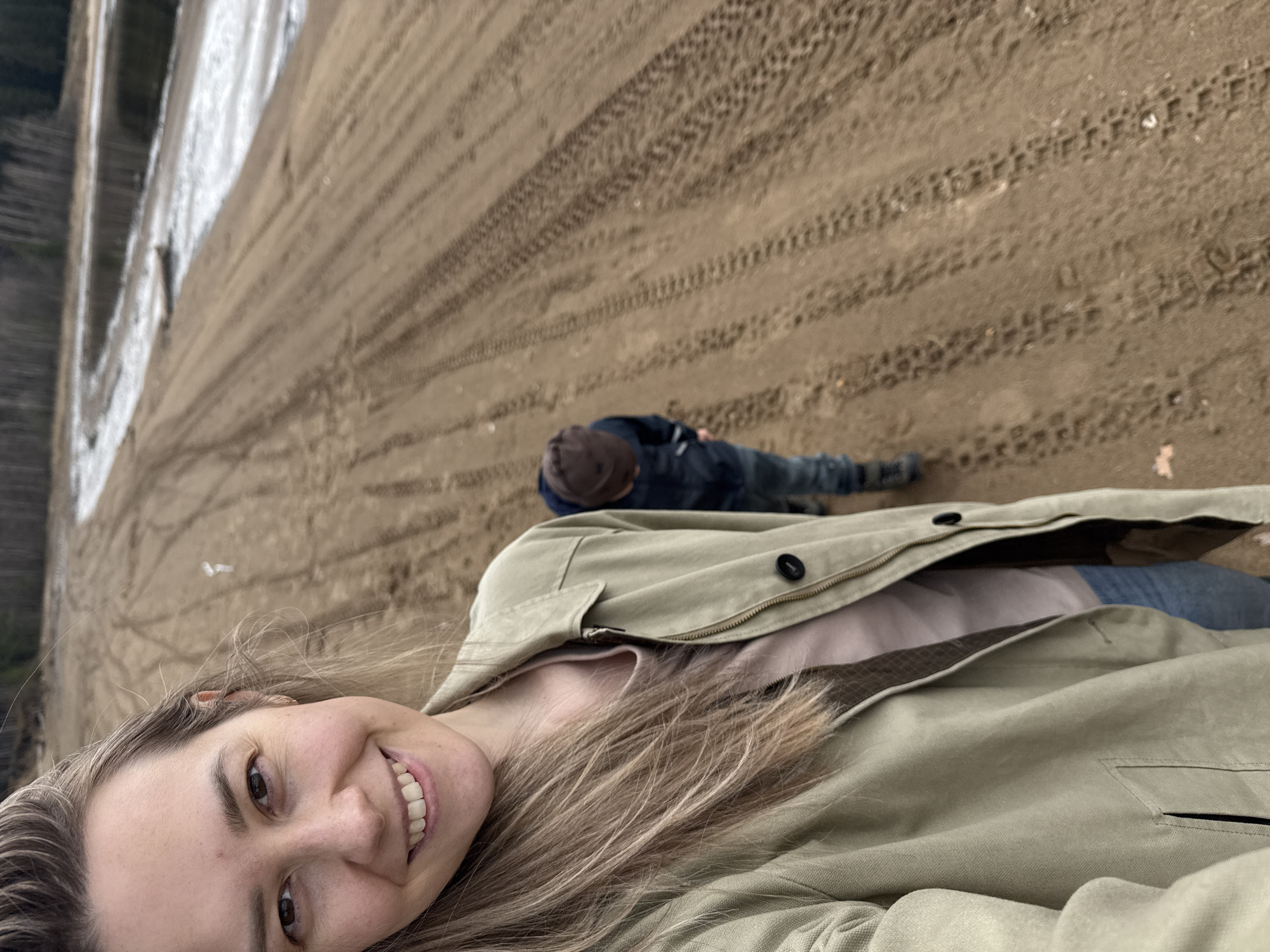 This selfie captures a smiling woman in the foreground with her long, light brown hair falling over her shoulder. She is wearing a light olive green or beige zippered jacket over a lighter top. In the mid-ground, a child or smaller person is seen lying or sitting on the sandy ground, wearing a dark jacket, jeans, and a dark cap. The setting is a sandy area, likely a beach or shoreline, characterized by numerous tire tracks imprinted on the sand. To the left, a body of water is visible, suggesting a river or lake, consistent with the Chaikovsky, Russia location context. The lighting appears overcast, indicating daytime. The woman's smile conveys a cheerful emotion. There is no visible text in the image. The scene depicts a casual outdoor outing.