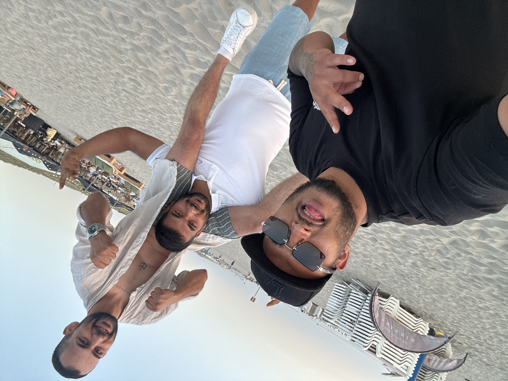 Three men are posing for a selfie on a sandy beach. The man in the foreground is wearing a black t-shirt and a black baseball cap, with sunglasses and his tongue sticking out. The second man is standing behind him, wearing a white polo shirt and light blue shorts. The third man is sitting on the second man's shoulders, wearing a light-colored open shirt and showing a tattoo on his chest. He is flexing his arms and wearing a watch. In the background, beach chairs are stacked, and a beach restaurant with people sitting at tables is visible. The scene appears to be at a beach location in The Hague, Netherlands, possibly Scheveningen. The sky is clear and the overall lighting suggests it is late afternoon or early evening.