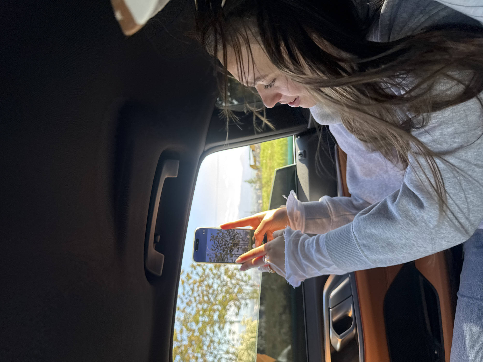 A young woman is positioned inside a car, holding a smartphone up to the open window to photograph a tree in bloom. The interior of the car has brown upholstery and a black grab handle. The woman wears a grey sweatshirt over a white blouse with ruffled cuffs and blue jeans. Through the window, the scene shows a tree with white blossoms against a backdrop of greenery. Based on the limited visual information, it is not possible to definitively confirm the location in Yerevan, Armenia.