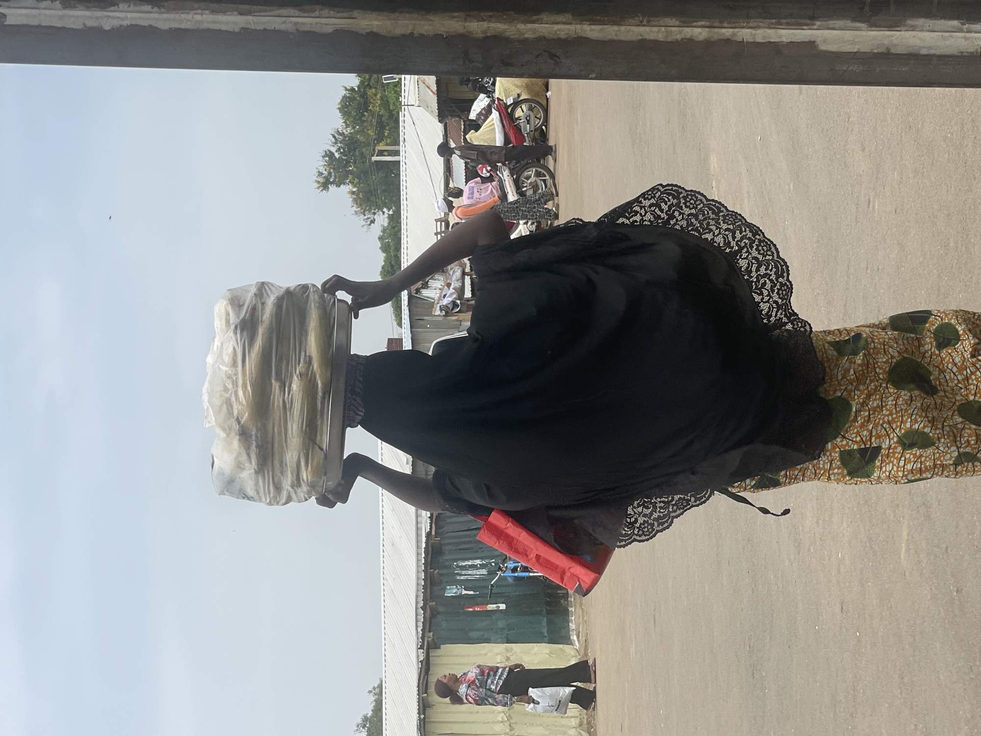 A woman carries a tray of bagged items on her head in Jos, Nigeria. She wears a black dress trimmed with lace over a patterned skirt. A red bag hangs from her arm. Behind her, there are motorbikes parked along a street lined with corrugated metal buildings. People are walking along the road. The sky is overcast.
