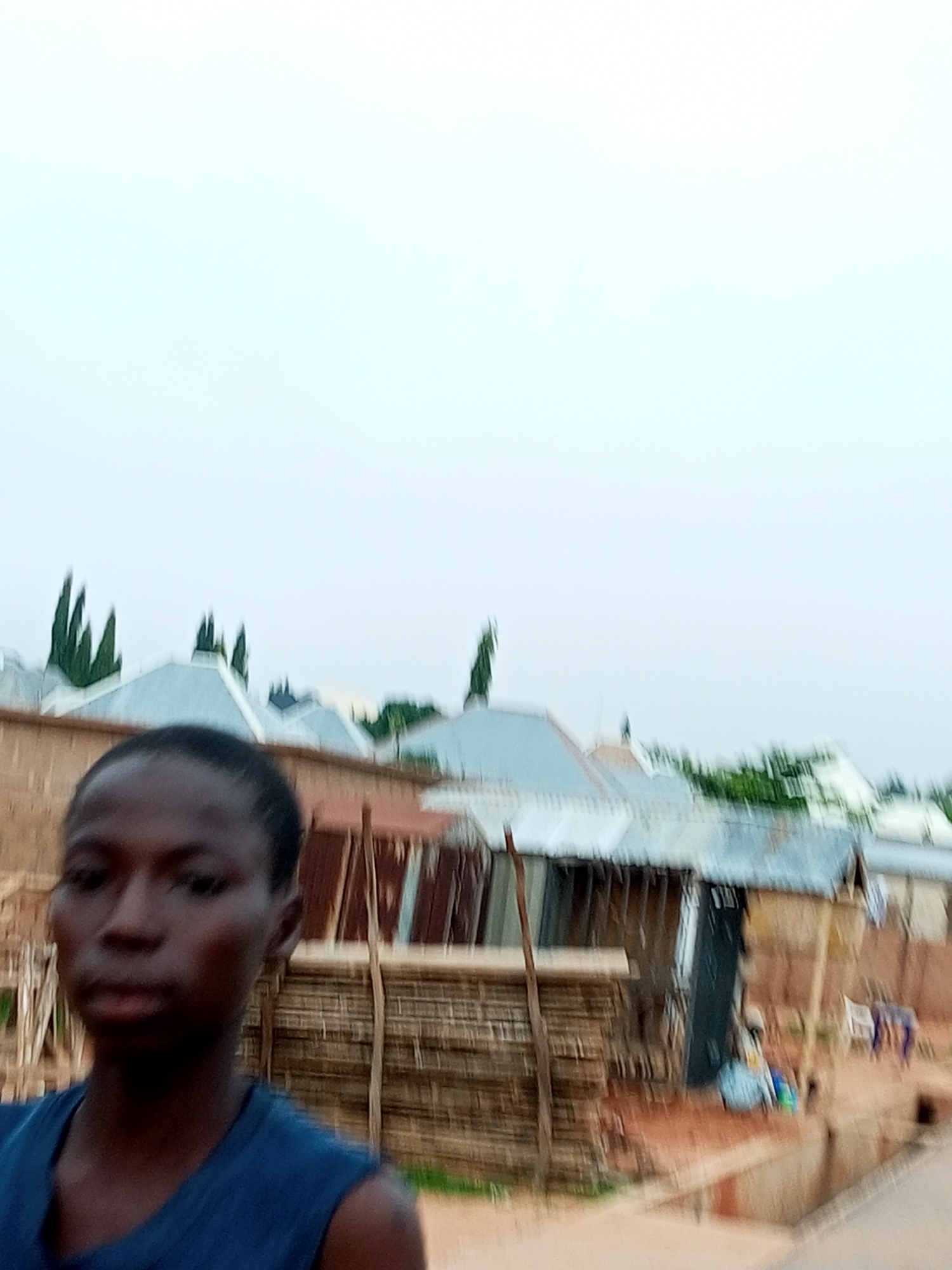 A young person with dark skin and dark hair is looking into the camera, wearing a blue sleeveless shirt. Behind them, there are several buildings with corrugated metal roofs, some appearing to be houses or small shops. The structures are a mix of materials, including what looks like bamboo or straw walls. In the background, there are tall, slender trees. The ground is made of reddish-brown earth. The overall impression is of a rural or semi-urban environment in Nigeria.