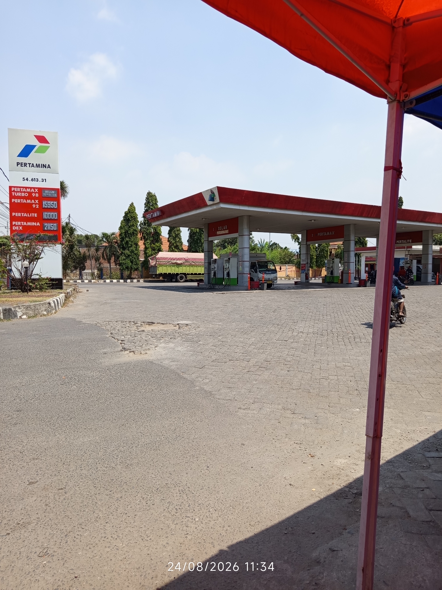 A Pertamina gas station is depicted in Canggu, Indonesia, on a sunny day. The gas station has a red roof and white pillars. Various fuel prices are displayed on a digital sign, including Pertamax Turbo at 98 and Pertamax 92 at 15,950. Pertalite is priced at 10,000, and Pertamina Dex at 21,150. A green truck is parked at one of the pumps, and a person on a motorcycle is riding away from the station. The ground is paved with cobblestones in front of the station and asphalt on the road leading to it, which shows signs of disrepair. Trees and buildings are visible in the background, along with a blue sky with scattered clouds. A red canopy extends from the right side of the frame, partially obscuring the view. The date and time stamp "24/08/2026 11:34" is visible in the bottom right corner.