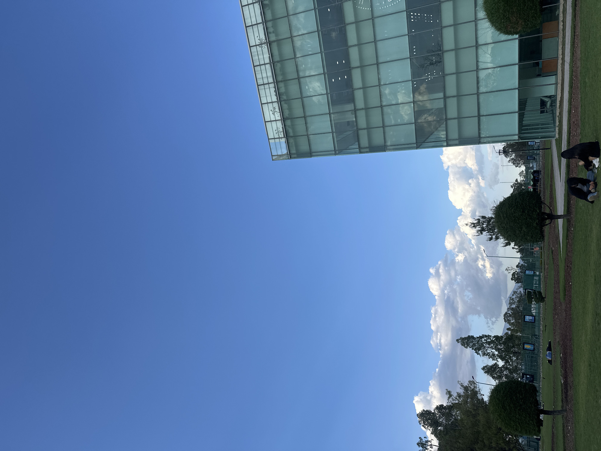 The image shows a cityscape with a clear blue sky taking up a large portion of the frame. On the right, the edge of a modern glass building is visible, reflecting the sky and clouds. Below the building, there is a grassy area with trees and bushes. Two individuals appear to be sitting or kneeling on the grass. In the background, there is a fence and some distant objects that might be street lights or other urban infrastructure. The location is Unknown, Unknown.