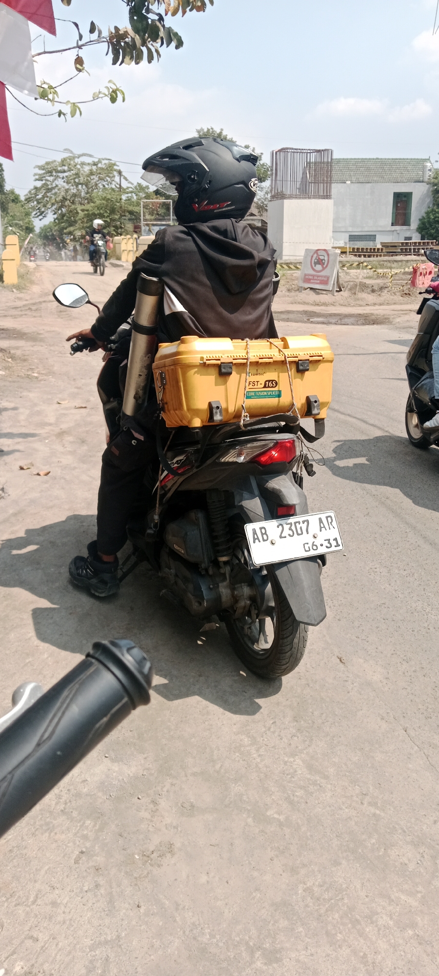 A person wearing a black helmet and black clothing is riding a black scooter on a dirt road in Purwomartani, Indonesia. The scooter has a yellow storage box attached to the back, which contains equipment labeled "FST-16S CORE FUSION SPLICER". A long cylindrical object is also attached vertically to the back of the scooter. The scooter's license plate reads "AB 2367 AR G6-31". In the background, there is construction work with a building under construction and a "NO PARKING" sign. Trees and blue sky are visible.