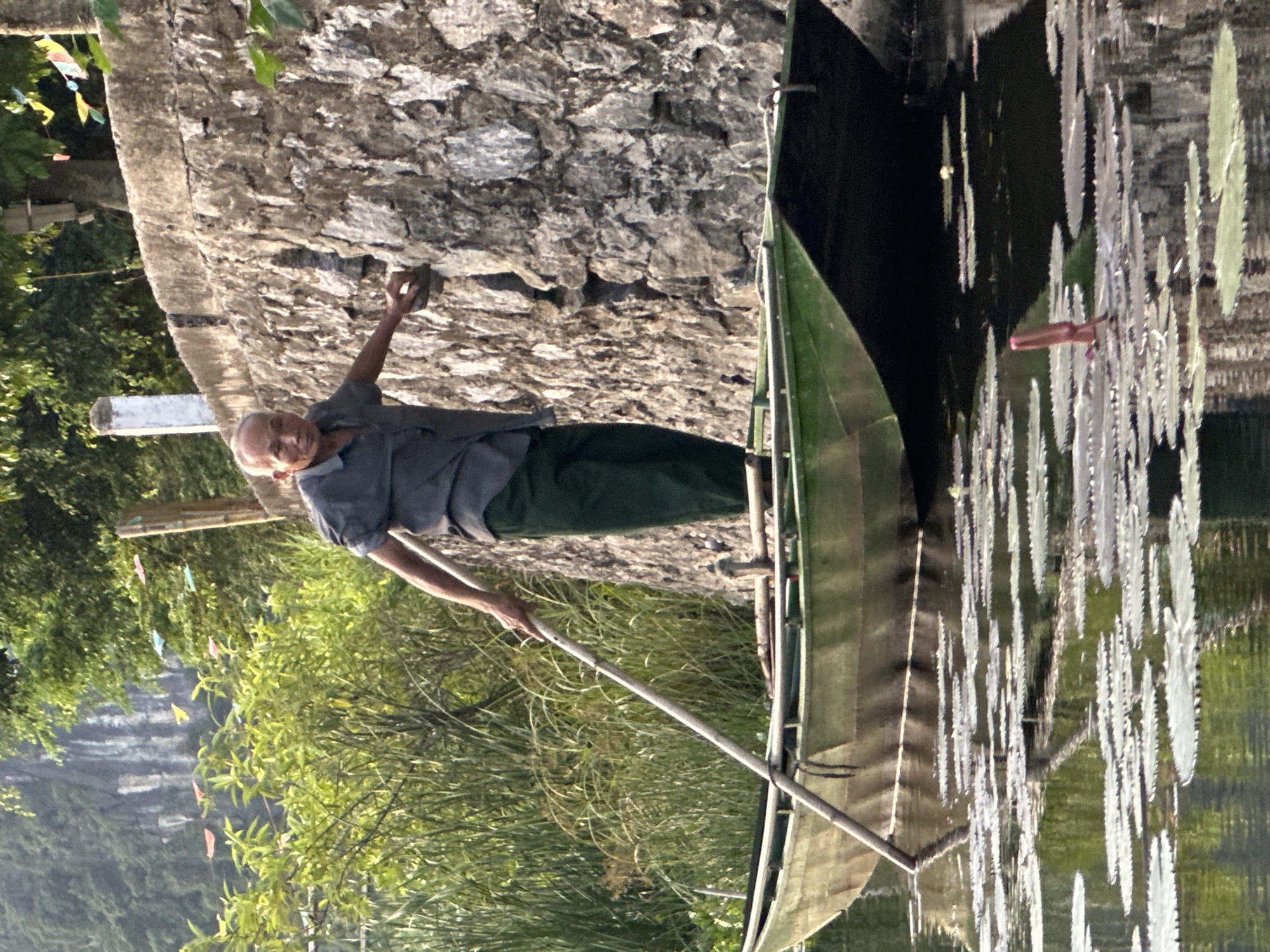 An elderly Vietnamese man, wearing a dark grey short-sleeved shirt and dark green pants, stands in a green and black rowboat. He is holding a long wooden pole, likely a oar or a pushing pole, which he is using to propel the boat through the water. The boat is filled with what appear to be water lilies, with some lily pads floating on the surface of the water. The man is facing towards the right side of the frame, looking at the viewer with a neutral expression. Behind him, a textured stone wall runs along the bank, overgrown with vegetation. In the background, lush green trees and a rocky mountainside are visible, suggesting a rural or natural setting. The overall lighting indicates it is daytime, with sunlight casting reflections on the water. This scene is located in Nam Hoa Lư Ward, Vietnam.