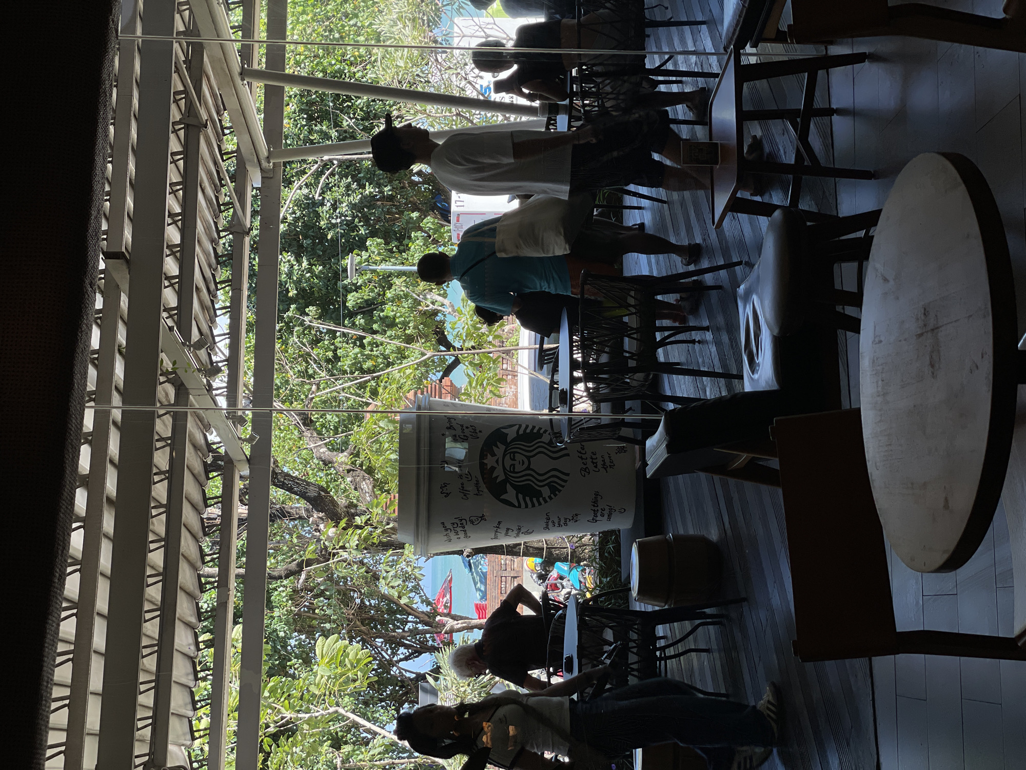The image depicts the outdoor seating area of a Starbucks in Kuta, Indonesia, on a sunny day. The scene is viewed through a glass-paneled enclosure, with a roof structure overhead supported by metal beams. Lush green trees and the ocean are visible in the background, with beach umbrellas indicating proximity to the coast. Inside the seating area, several people are present. In the foreground, a woman with dark hair pulled back is walking away from the camera, wearing a white t-shirt and dark pants. An older man with white hair is seated at a table, wearing a dark t-shirt and shorts. Further back, a man in a blue t-shirt and orange shorts carries a large white tote bag, walking away from the camera. Another man in a white t-shirt and dark shorts walks towards the right of the frame. Several tables and chairs, some made of dark wood and others with woven seats, are arranged on a wooden deck floor. A prominent feature in the middle ground is a large, oversized Starbucks cup replica, adorned with various messages and the iconic Starbucks logo. In the distance, other patrons are seated at tables, and a street lamp is visible amongst the trees. The overall impression is that of a casual, open-air cafe environment.