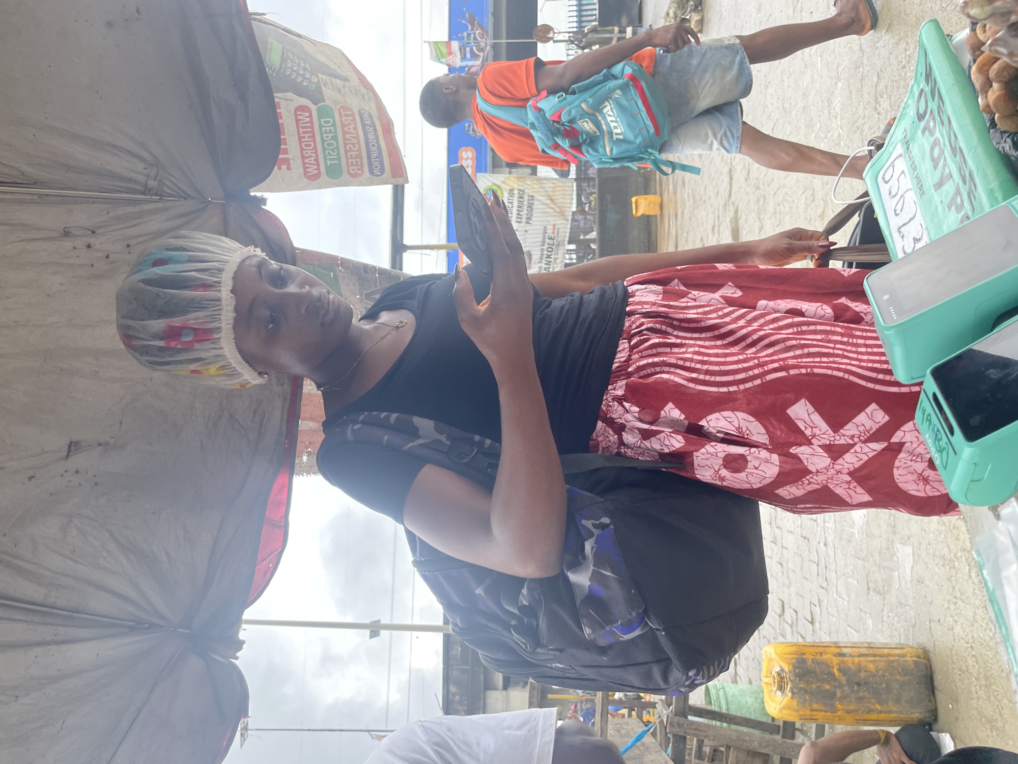 In Lekki, Nigeria, a young woman wearing a floral shower cap and a black t-shirt stands under a makeshift canopy. She is holding a smartphone and looking down at it. A large, dark-colored backpack with blue and grey camouflage patterns is slung over her left shoulder. She is also wearing a red skirt with a white geometric pattern. To her right, a young man in an orange t-shirt and denim shorts walks away from the camera, carrying a turquoise backpack. In the foreground, two green point-of-sale devices are visible, along with what appears to be a pile of coconuts. A large yellow plastic container and a green plastic container are also present in the scene, as are various market stalls and signage in the background. The ground appears wet, suggesting recent rain.