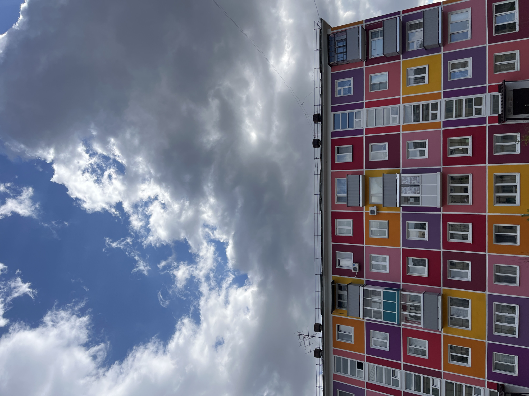 The image shows a colorful apartment building in Zelenogorsk, Russia, viewed from below. The building's facade is composed of square sections painted in purple, yellow, pink, red, and orange hues, each containing one or more windows. A metal ladder and utility fixtures are attached to the side of the building. The sky is visible on the left, featuring a mix of dark storm clouds and patches of blue sky. A wire runs across the top of the building to the left.
