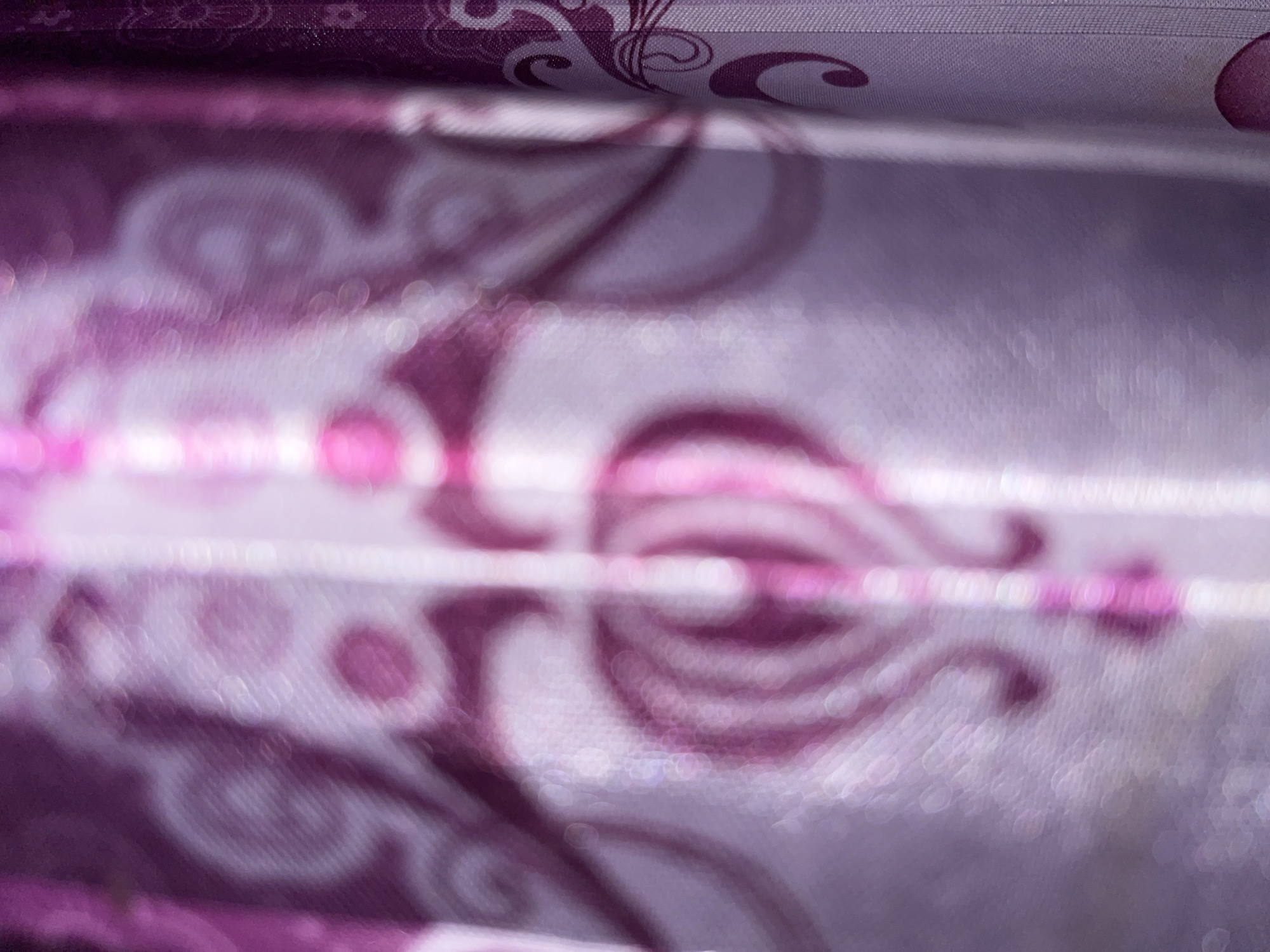 The image displays a close-up, out-of-focus view of a patterned curtain in shades of purple and pink. The fabric features swirling floral or paisley designs, with some circular bokeh effects from light sources. The curtain appears to be made of a sheer or semi-sheer material, allowing some light to pass through. The scene is indoors, suggesting a domestic setting. There are no people or identifiable objects from Buleleng, Indonesia, visible in this image.