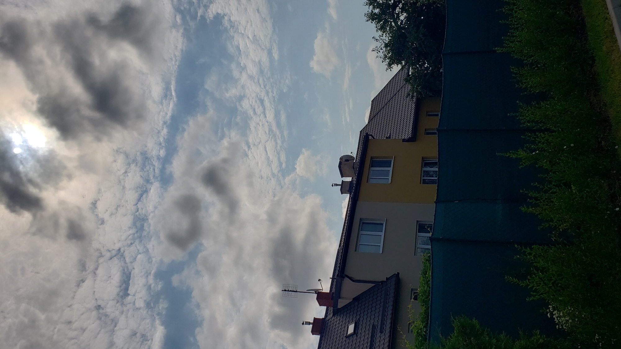 This is a vertical, outdoor, medium shot image taken on a cloudy day with the sun partially obscured by clouds in Prešov, Slovakia. The scene features the roofs and upper floors of several attached residential buildings. The buildings have dark brown tiled roofs, and the facades of the buildings are primarily cream and yellow. Windows on the upper floors are visible, some with white frames. Antennas and satellite dishes are mounted on some of the roofs. In the foreground, a dark green mesh fence runs horizontally across the image, behind which lush green vegetation and some small white flowers are growing. The sky is a mix of blue with scattered white and dark gray clouds.