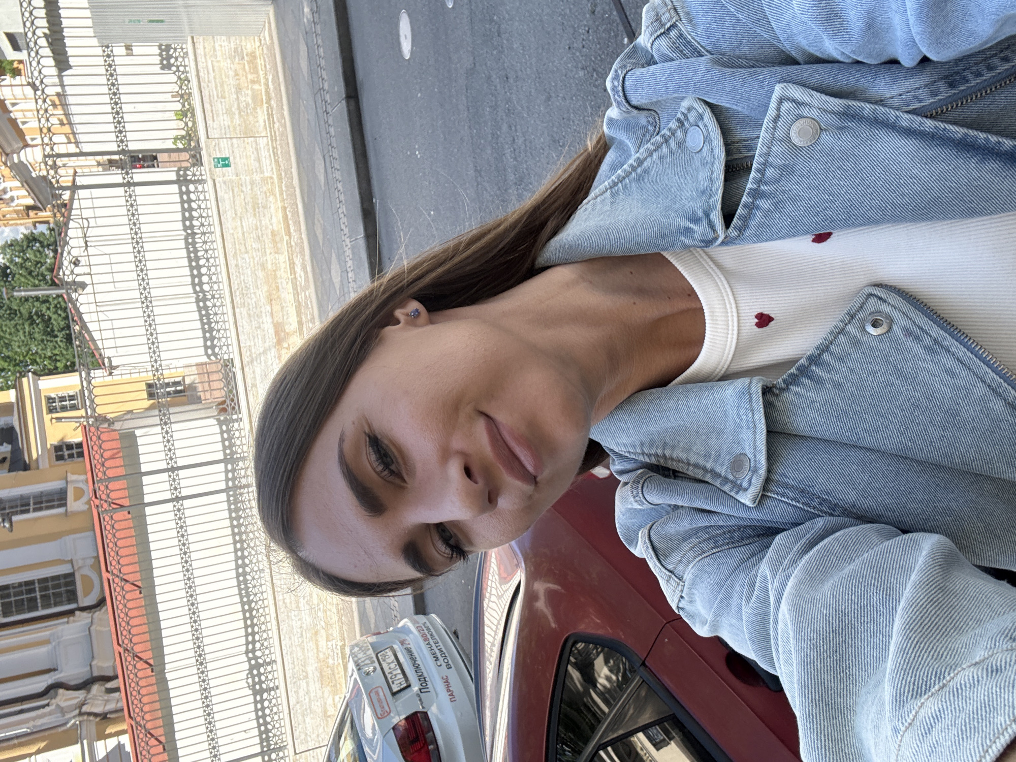 The image is a selfie taken by a woman in a denim jacket, with a white shirt featuring small red hearts. She is standing outdoors on a paved street, with two cars visible behind her: a red car on the left and a white car on the right. In the background, there is a building with yellow and white detailing and an ornate metal fence. The architecture and style of the building suggest the location is likely in the historic center of Saint Petersburg, Russia, possibly near a palace or a museum, given the ornate fence and building design. The lighting indicates it is daytime, with sunlight casting shadows on the ground. The woman is looking at the camera and smiling.