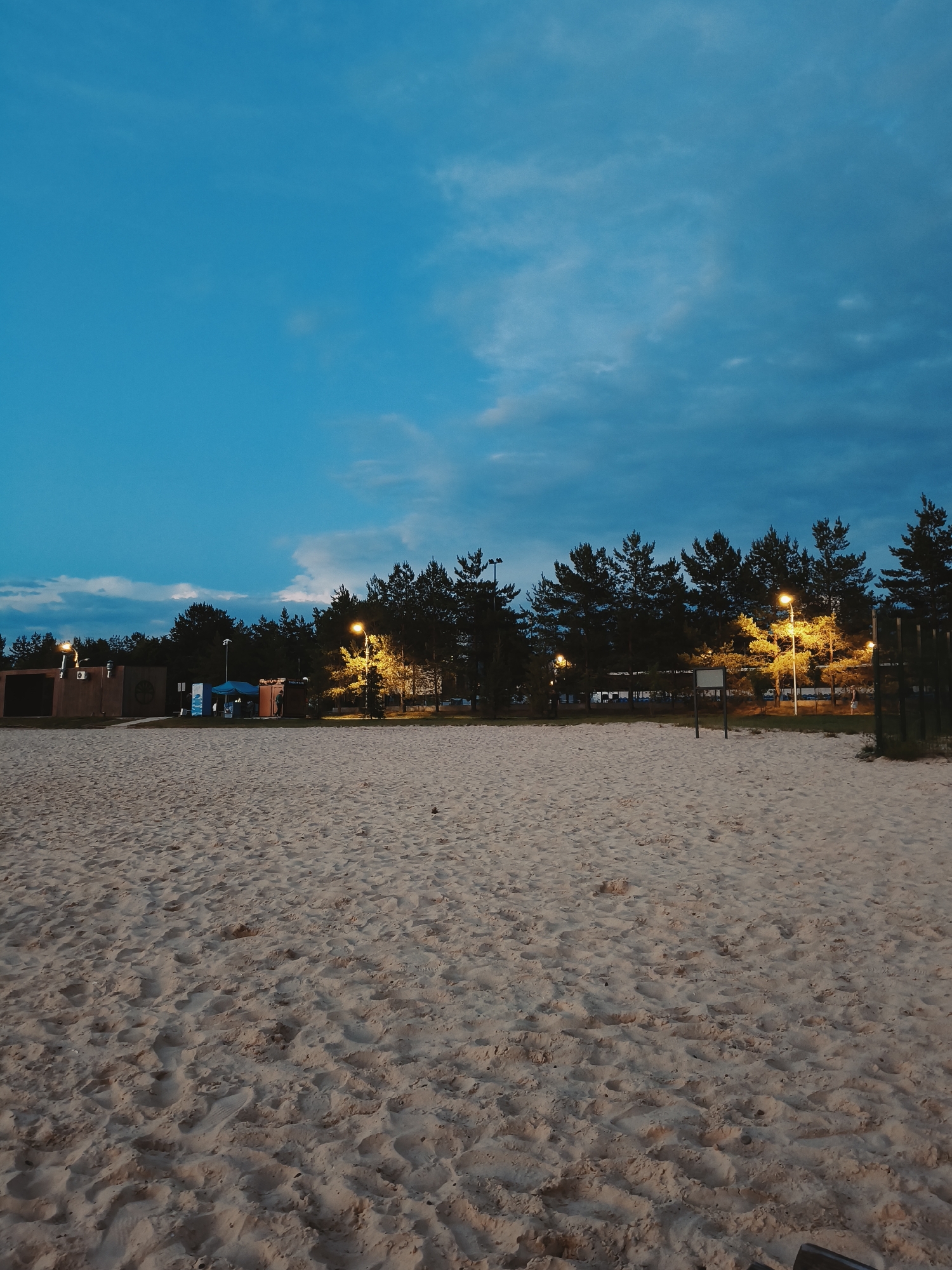 The image depicts a sandy beach at dusk, with trees forming a dark silhouette against a twilight sky. Several lampposts illuminate the scene with a warm, yellow glow, casting light on the sand and trees. In the background, there are several small buildings or structures, some with lighting, and a dark fence. The sand is textured with footprints and undulations, suggesting it is a public area. The sky is a deep blue, with some lighter clouds visible near the horizon. The overall atmosphere is serene and quiet, characteristic of a beach at the end of the day. This scene is located at the "Ak-Chishma" beach on the shores of Lake Shaitankul in Ufa, Russia.