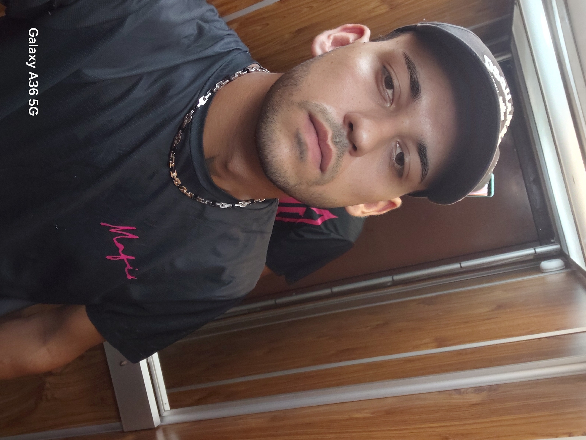 A young man wearing a black t-shirt with pink text that appears to say "Mafia" and a grey baseball cap takes a selfie in an elevator. The elevator has wood-paneled walls and a metal door frame. In the background, another person wearing a similar black shirt with pink lettering is visible, partially obscured. The text "Galaxy A36 5G" is present in the lower left corner of the image, indicating it was taken with a Samsung Galaxy A36 5G phone. The scene appears to be indoors, possibly in a building within the Municipal Unit of Aegio, Greece, as suggested by the user's prompt, though this cannot be definitively confirmed from the image alone.