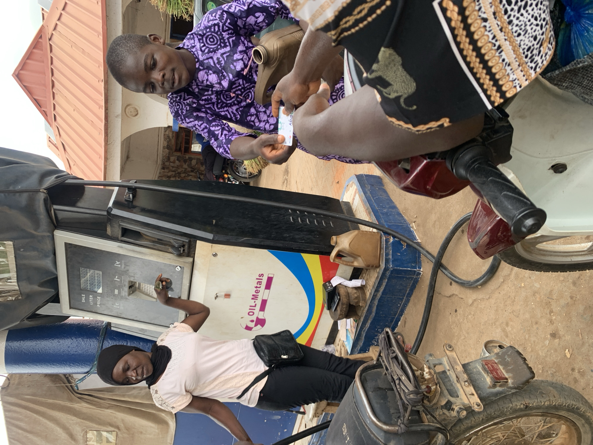 The image shows a scene at a gas station in Akwanga, Nigeria. A woman wearing a headscarf is operating a gas pump. Two men are interacting near a motorcycle; one man in a purple patterned shirt hands a credit card to the other. A small gas container sits near the pump. The gas station has an “Oil-Metals” logo. The surrounding environment appears to be a typical street scene with a building visible in the background.
